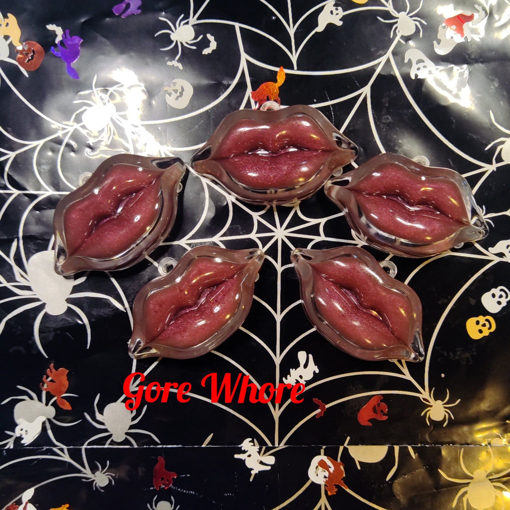 Lip-shaped resin pieces with red glitter, clear outline, on black spiderweb background with "Gore Whore" text