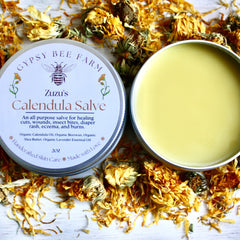 Calendula Salve, yellow salve in metal tin, surrounded by dried orange flower petals, Zuzu's brand visible