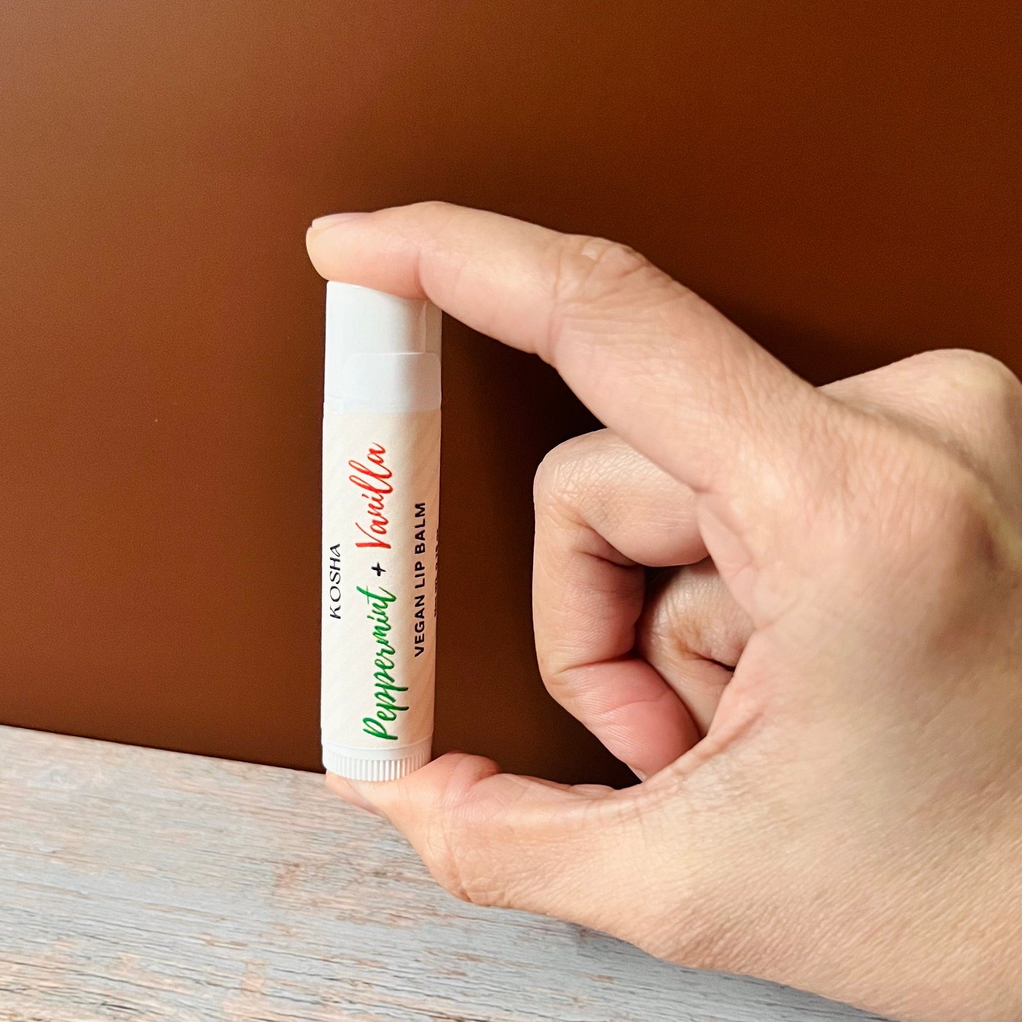 Lip balm tube, white with green text, held in hand, KOSHA brand visible.