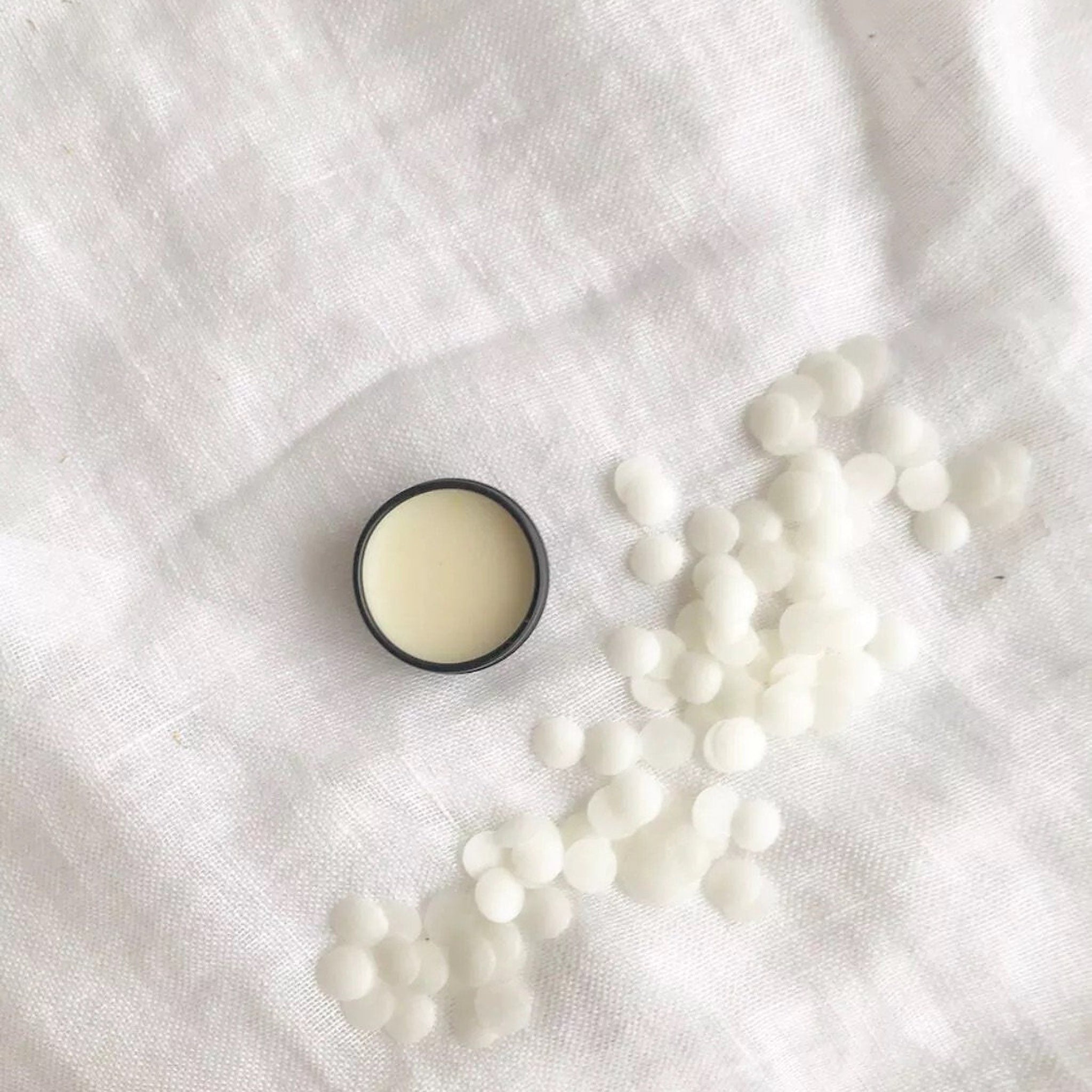 Lip balm in black container with light yellow balm on white linen background and white wax beads