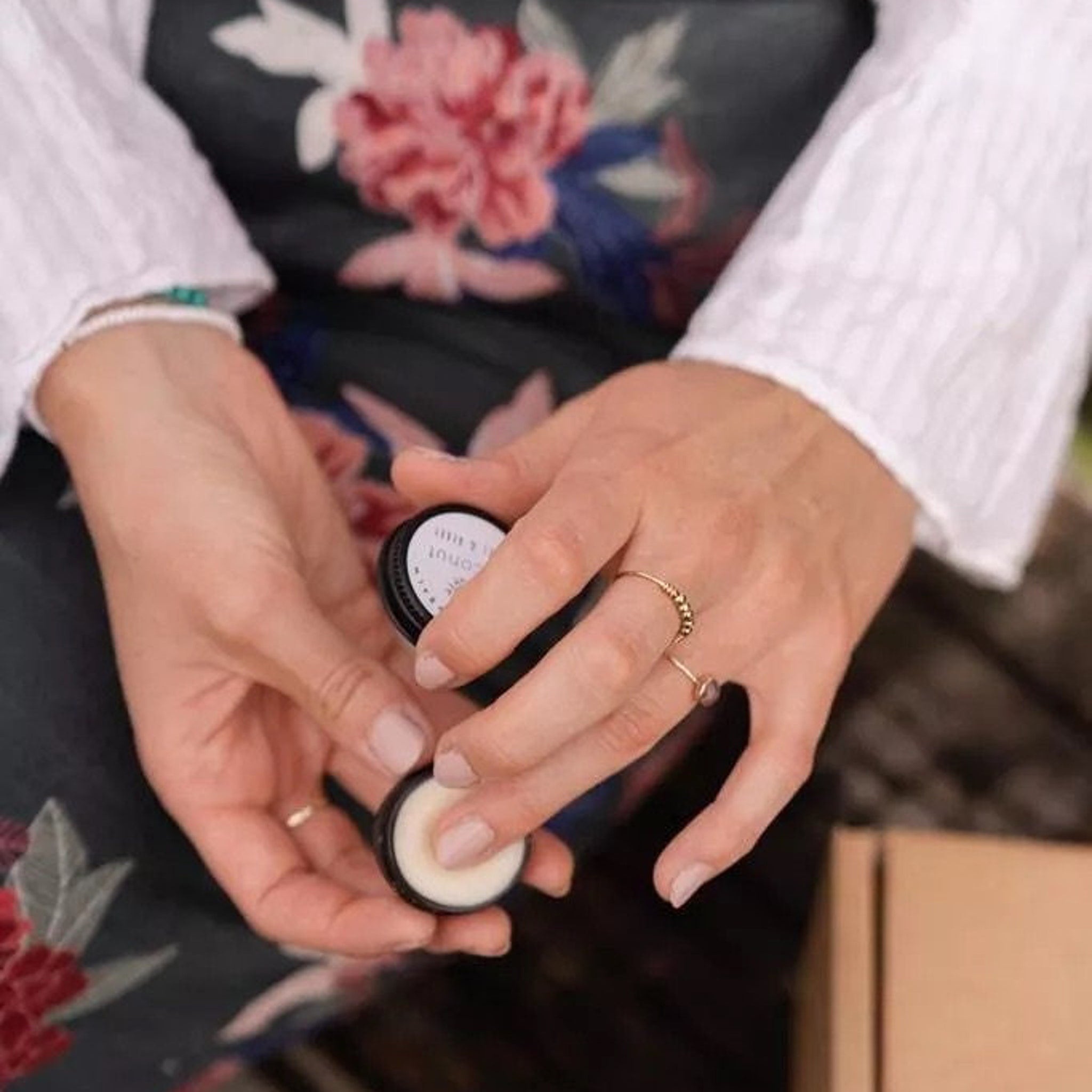 Lip balm in black container, hands holding, neutral nails, gold ring, floral background