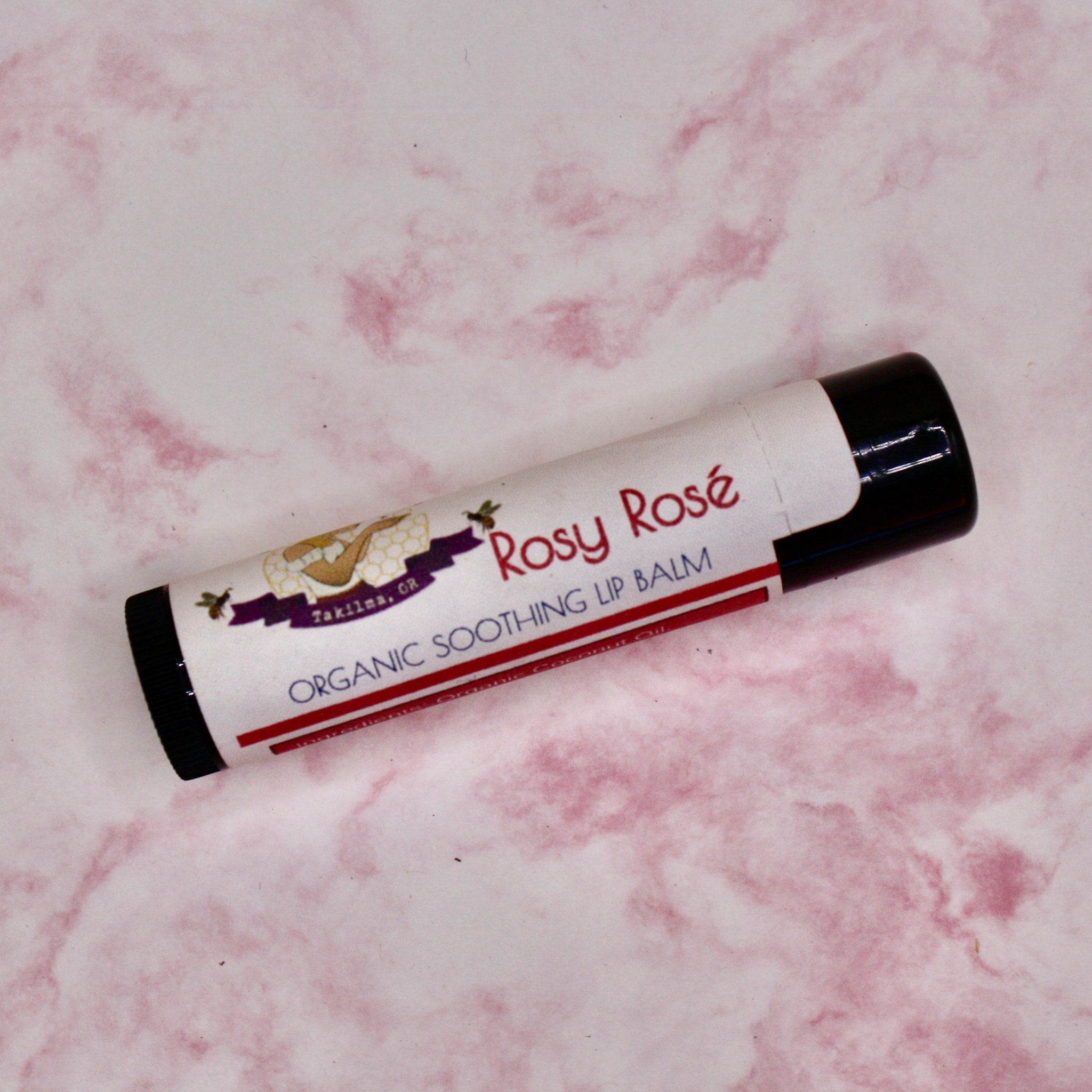 Lip balm tube with white label, black cap, and "Rosy Rosé" text on pink marble background