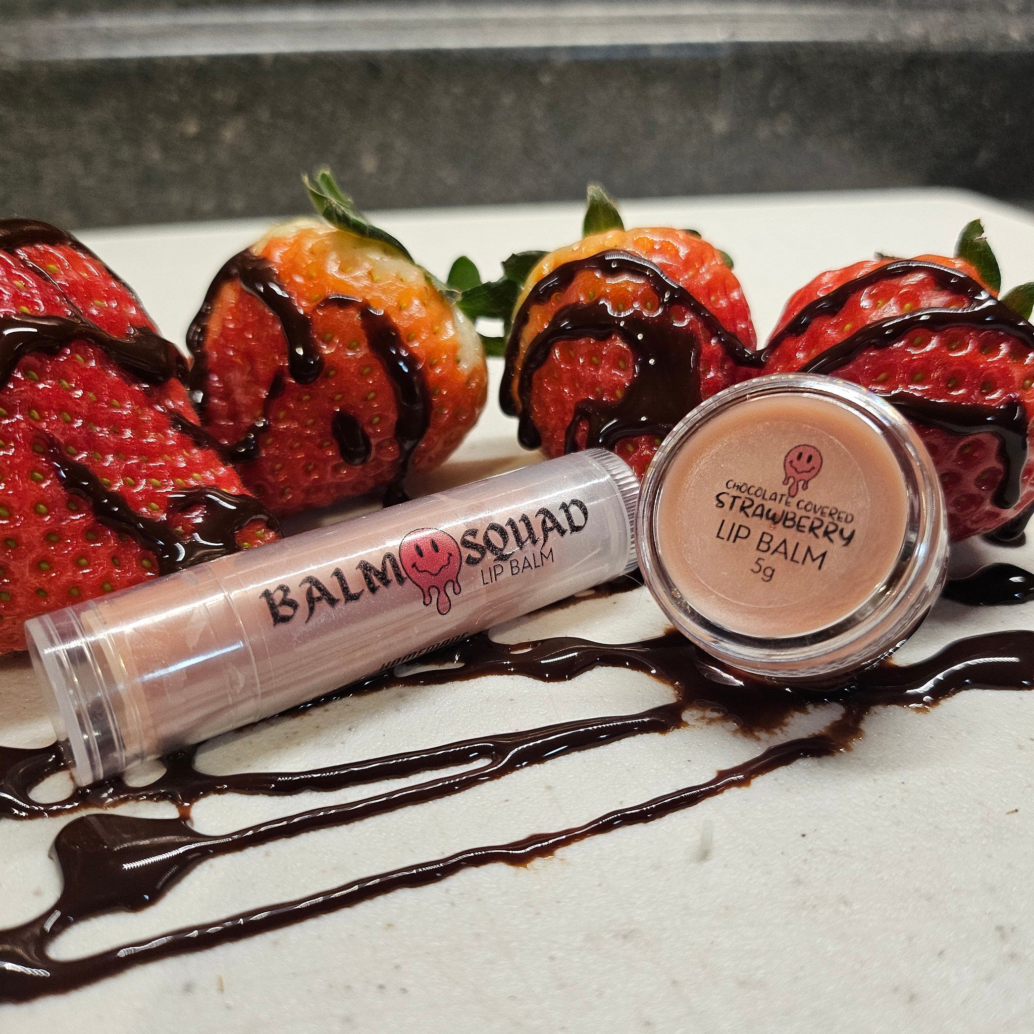 Lip balm with clear tube, pink balm, and "BALM SQUAD" text, next to chocolate-covered strawberries