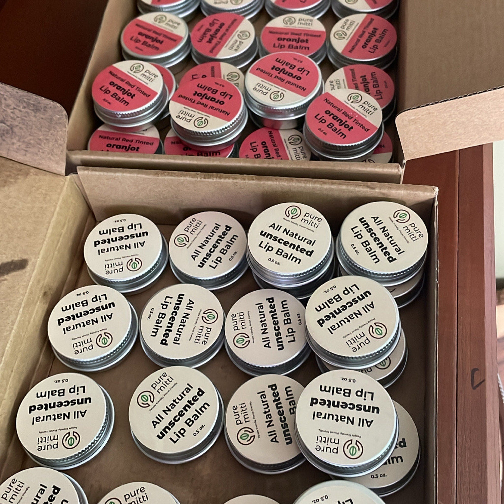 Lip balm tins with white labels, silver lids, and Pure Mitti branding, in boxes