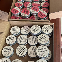 Lip balm tins with white labels, silver lids, and Pure Mitti branding, in boxes