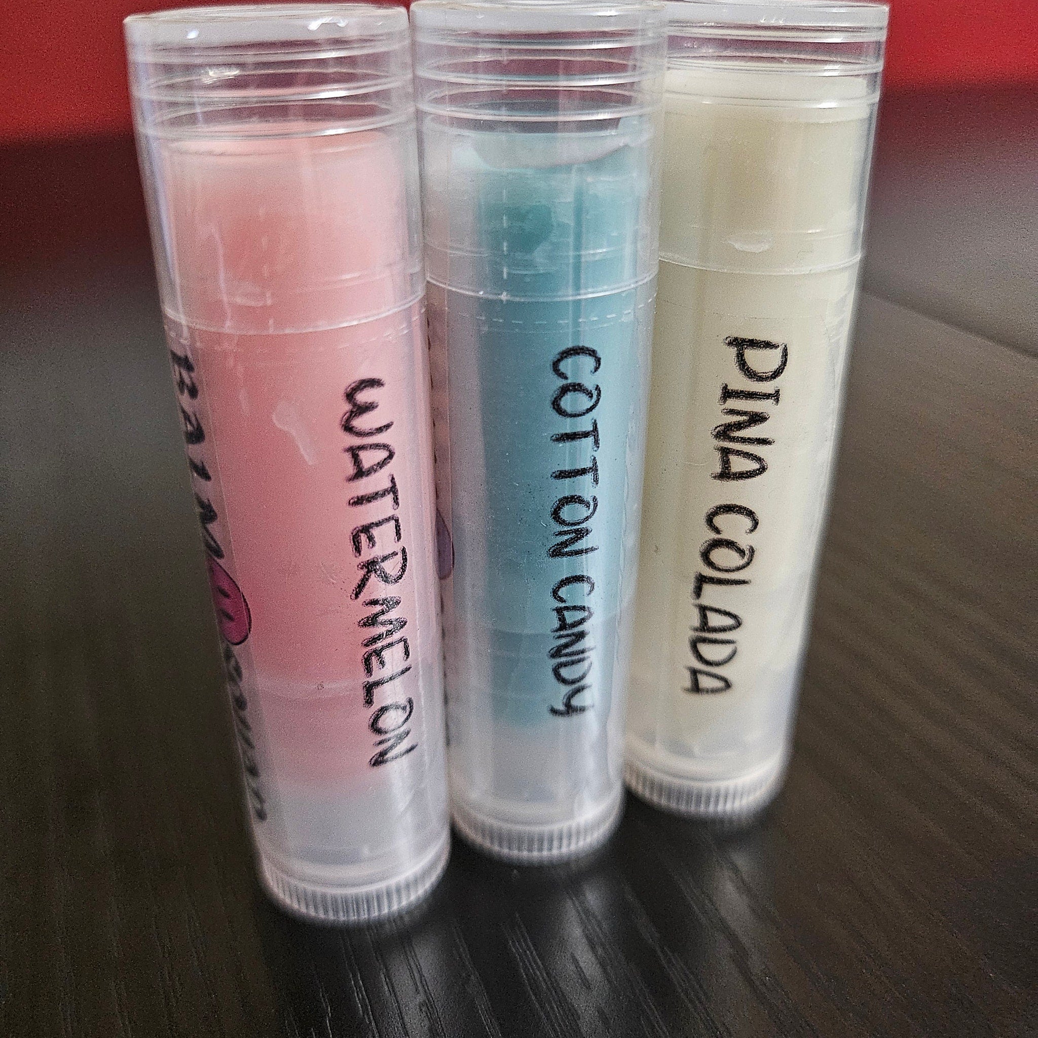 Lip balm tubes with pink, blue, and off-white color, with handwritten labels WATERMELON, COTTON CANDY, PINA COLADA