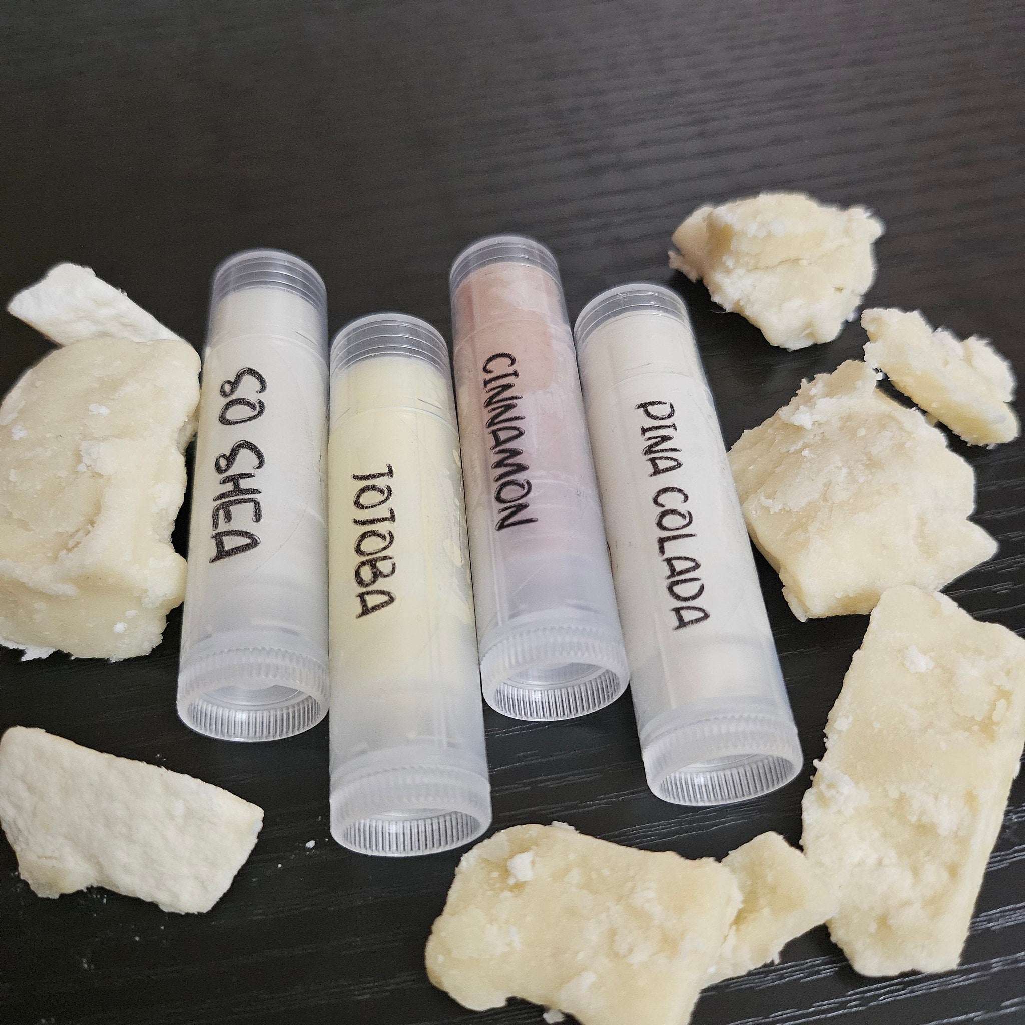 Lip balms in clear tubes with handwritten labels and white shea butter chunks on black surface