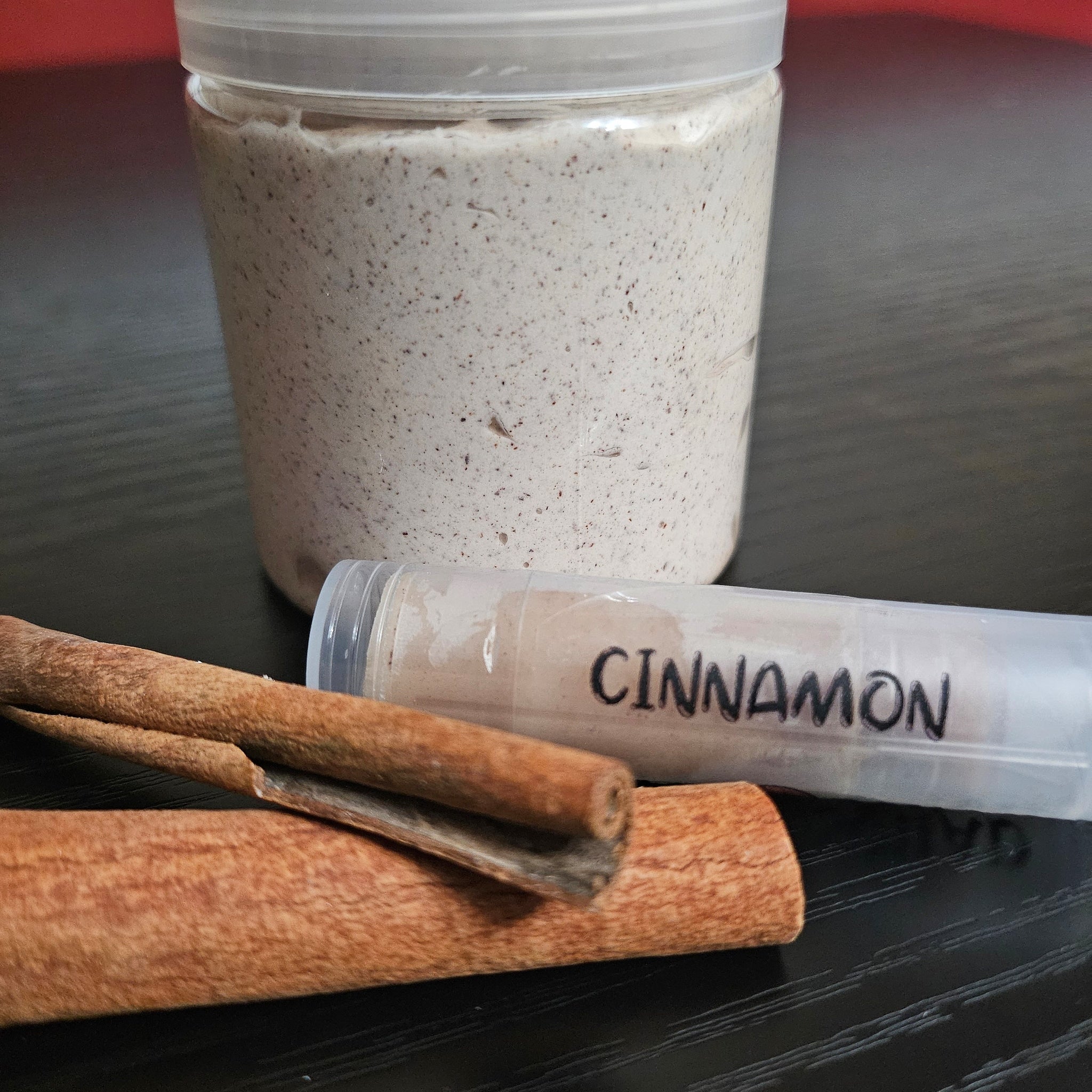 Cinnamon sticks with brown bark and "CINNAMON" text on a clear tube, next to a jar of speckled cream substance