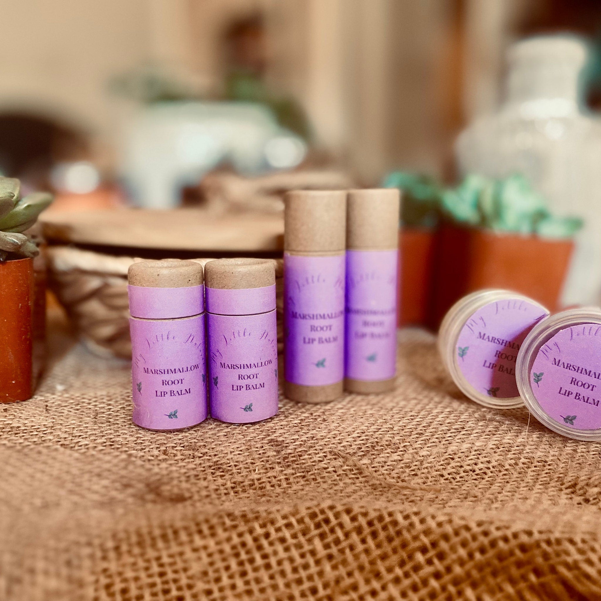 Lip balm tubes and jars in purple and brown, with text and small leaf design on burlap surface