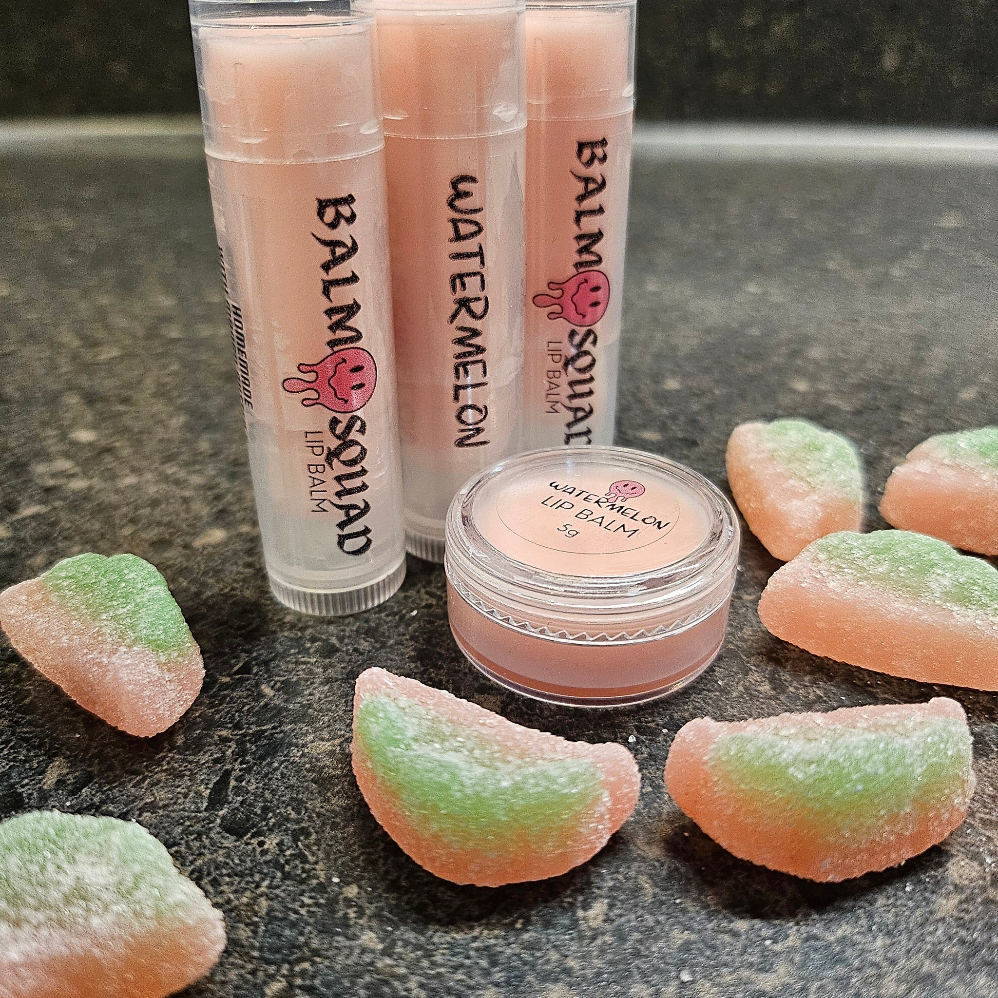 Lip balm tubes with pink balm and "Balm Squad" text, plus watermelon candy slices on dark surface