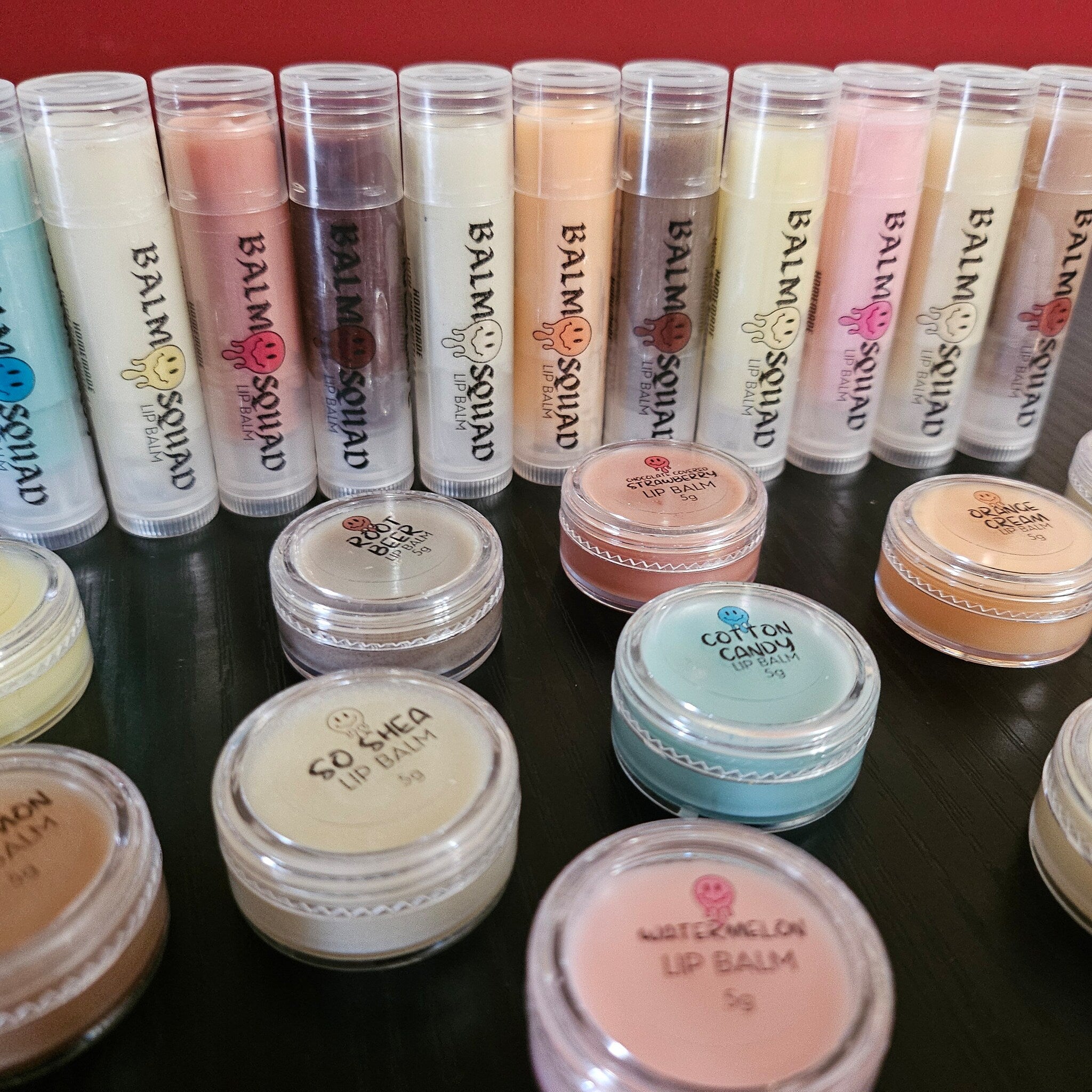 Lip balm tubes and jars in various colors, with "BALM SQUAD" text and smiley face logo visible