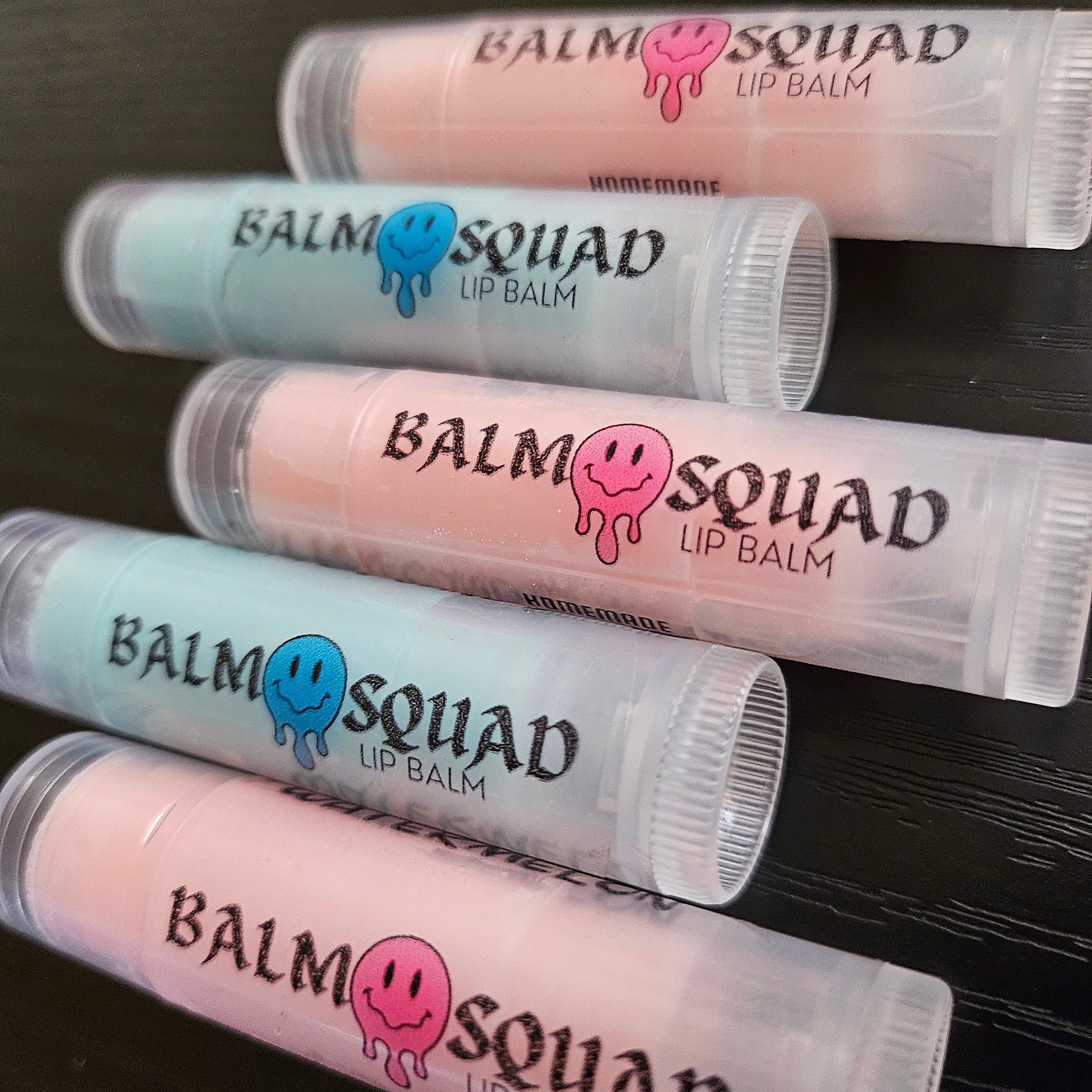 Lip balm tubes in pink, blue, and clear with "BALM SQUAD LIP BALM" text and smiley face logo visible