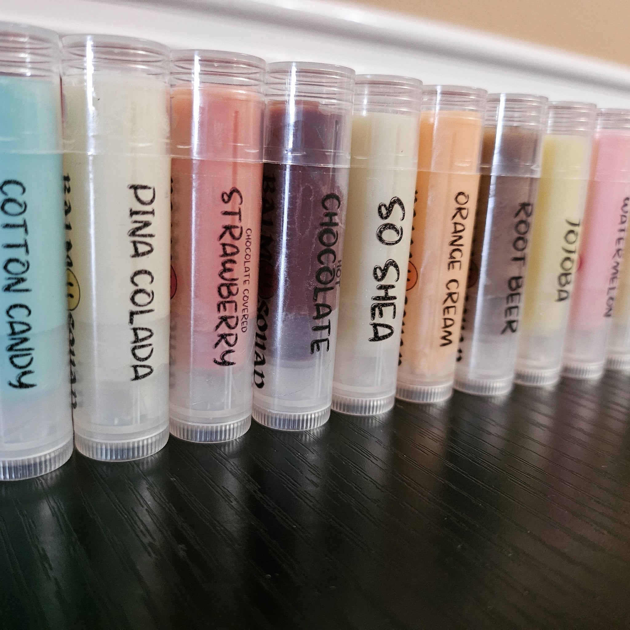 Lip balm tubes in various colors with handwritten flavor labels, plastic containers
