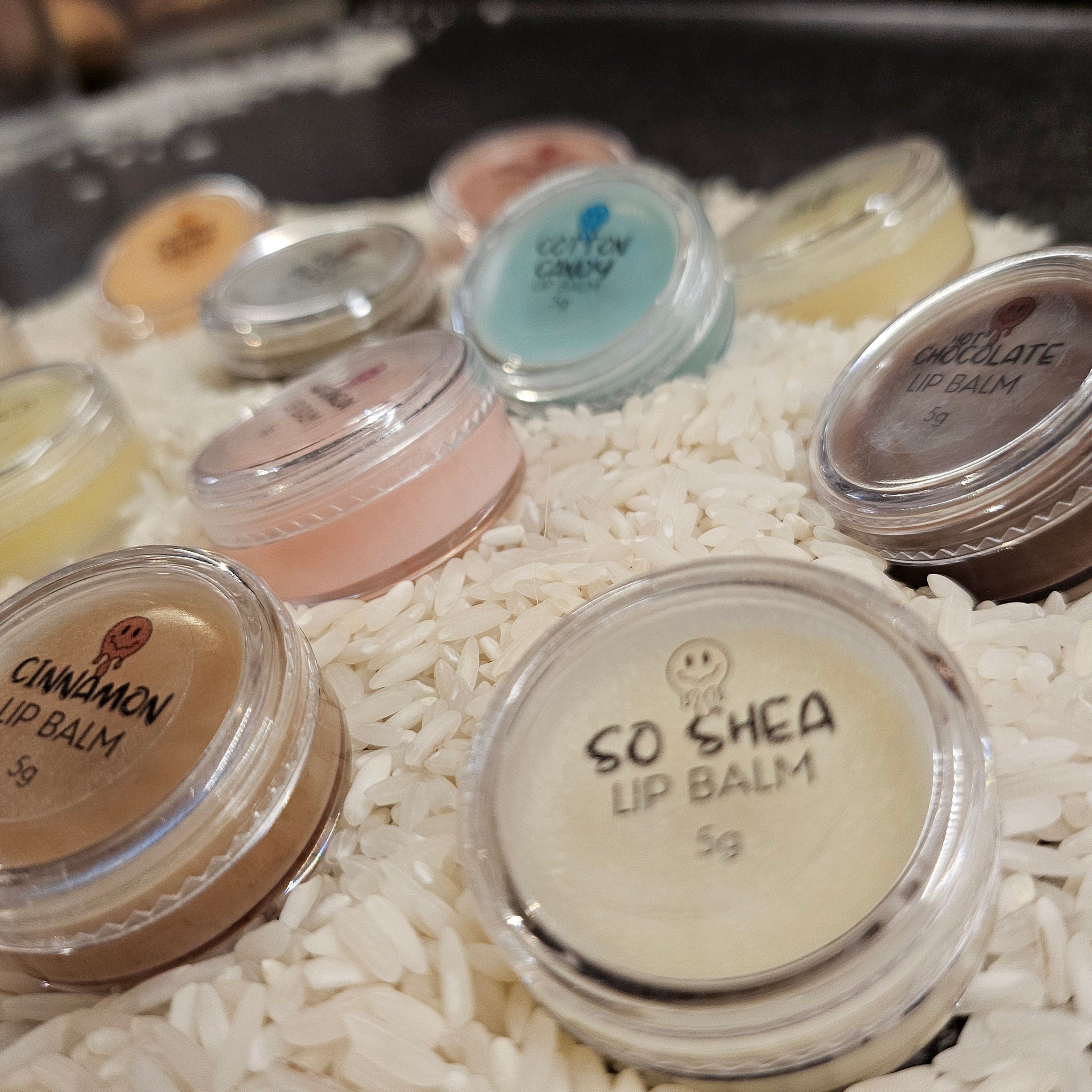 Lip balm jars in various colors, clear plastic containers, resting on white rice, with visible text labels
