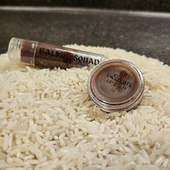 Lip balm in brown and clear tube and jar, on white rice, "Balm Squad" text visible