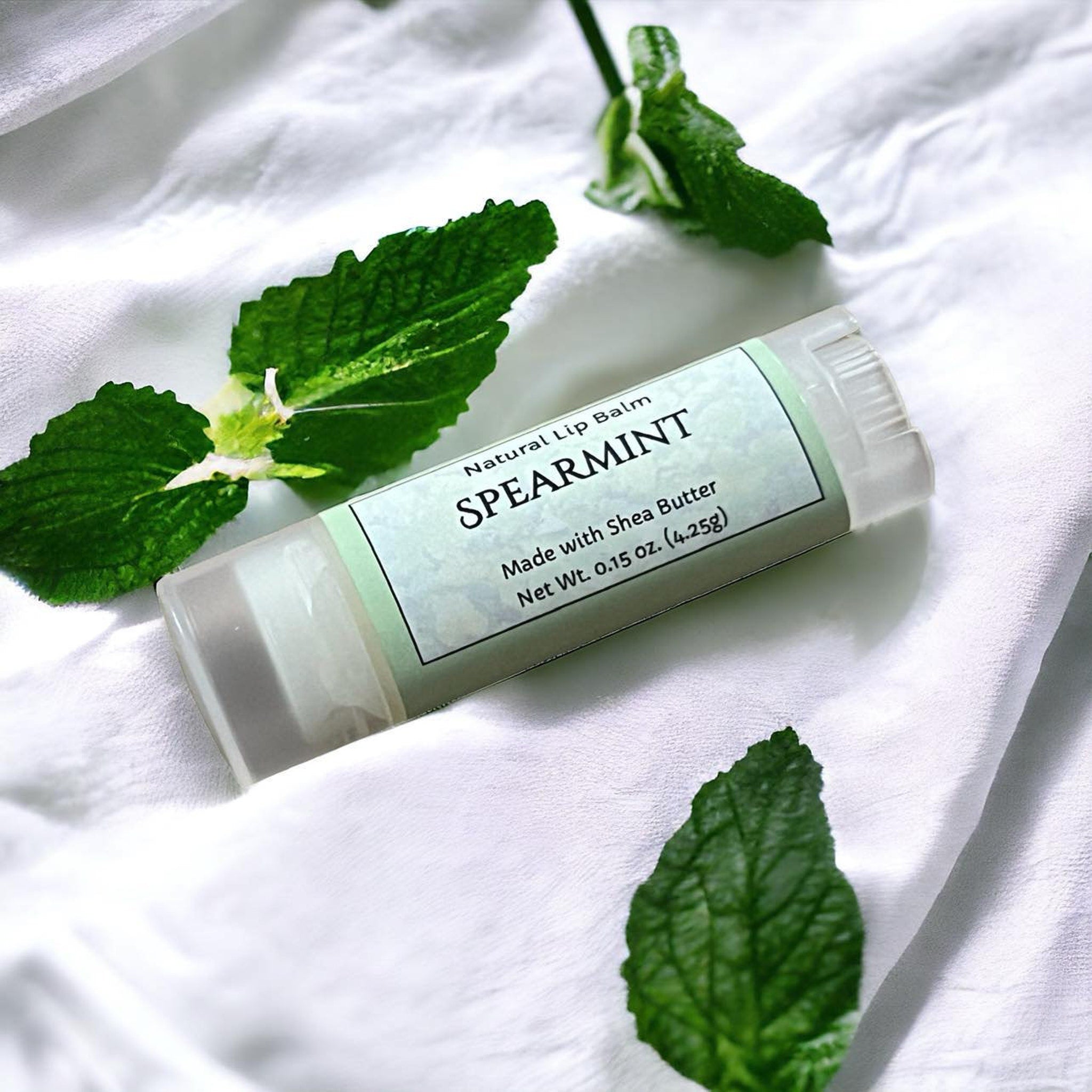 Lip balm tube with SPEARMINT label, clear plastic, white fabric background, and green mint leaves