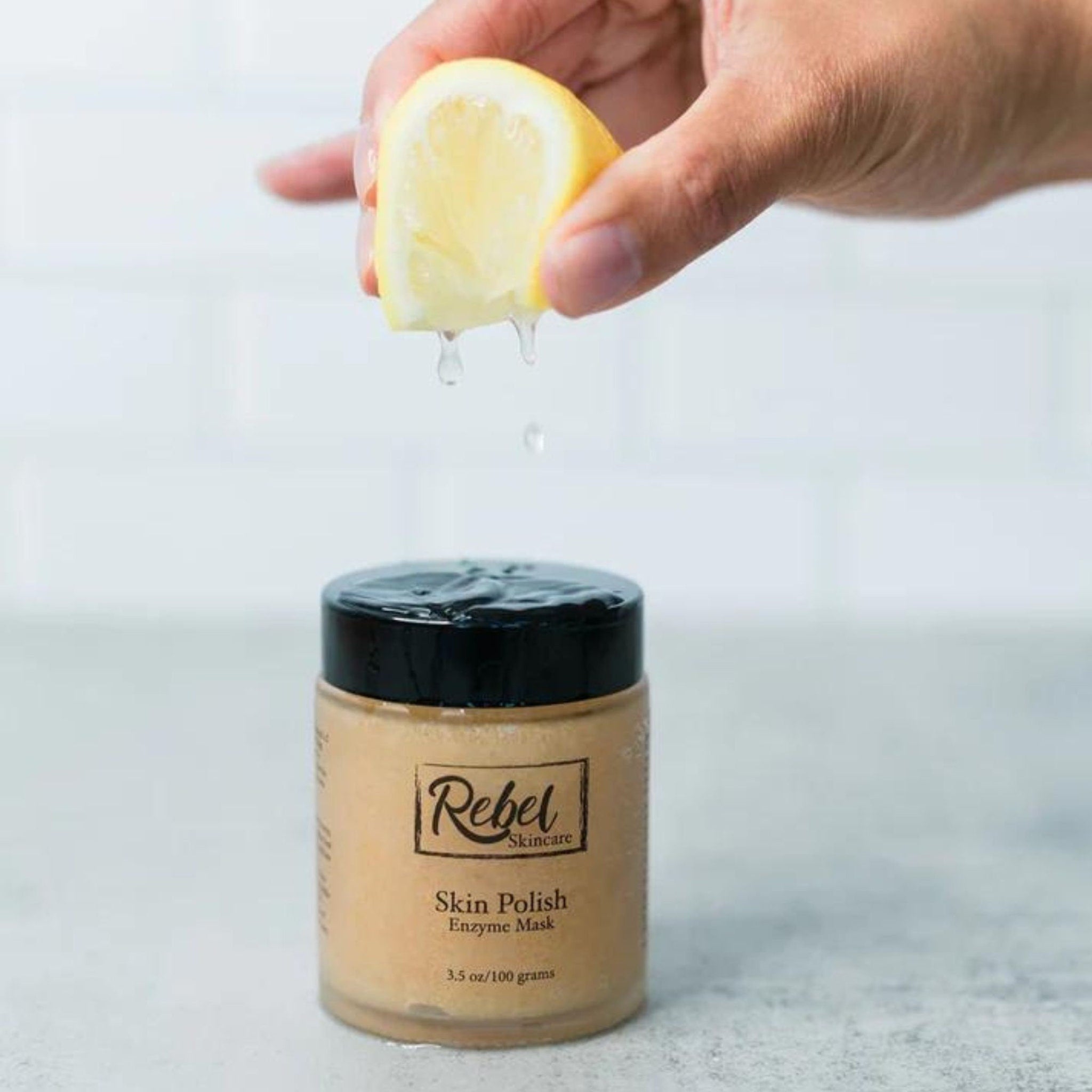 Skin polish jar with black lid, tan color, and Rebel Skincare label with lemon juice dripping over it