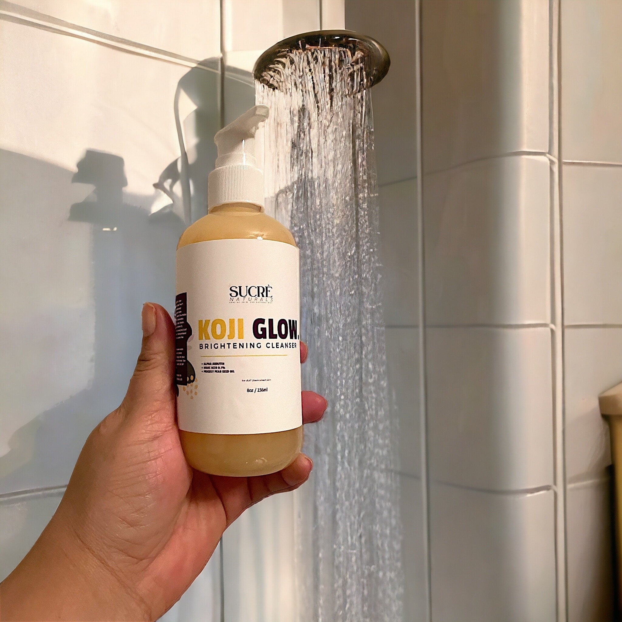 Koji Glow brightening cleanser in a yellow bottle, held in hand under shower water