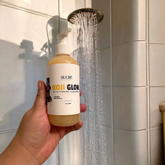 Koji Glow brightening cleanser in a yellow bottle, held in hand under shower water