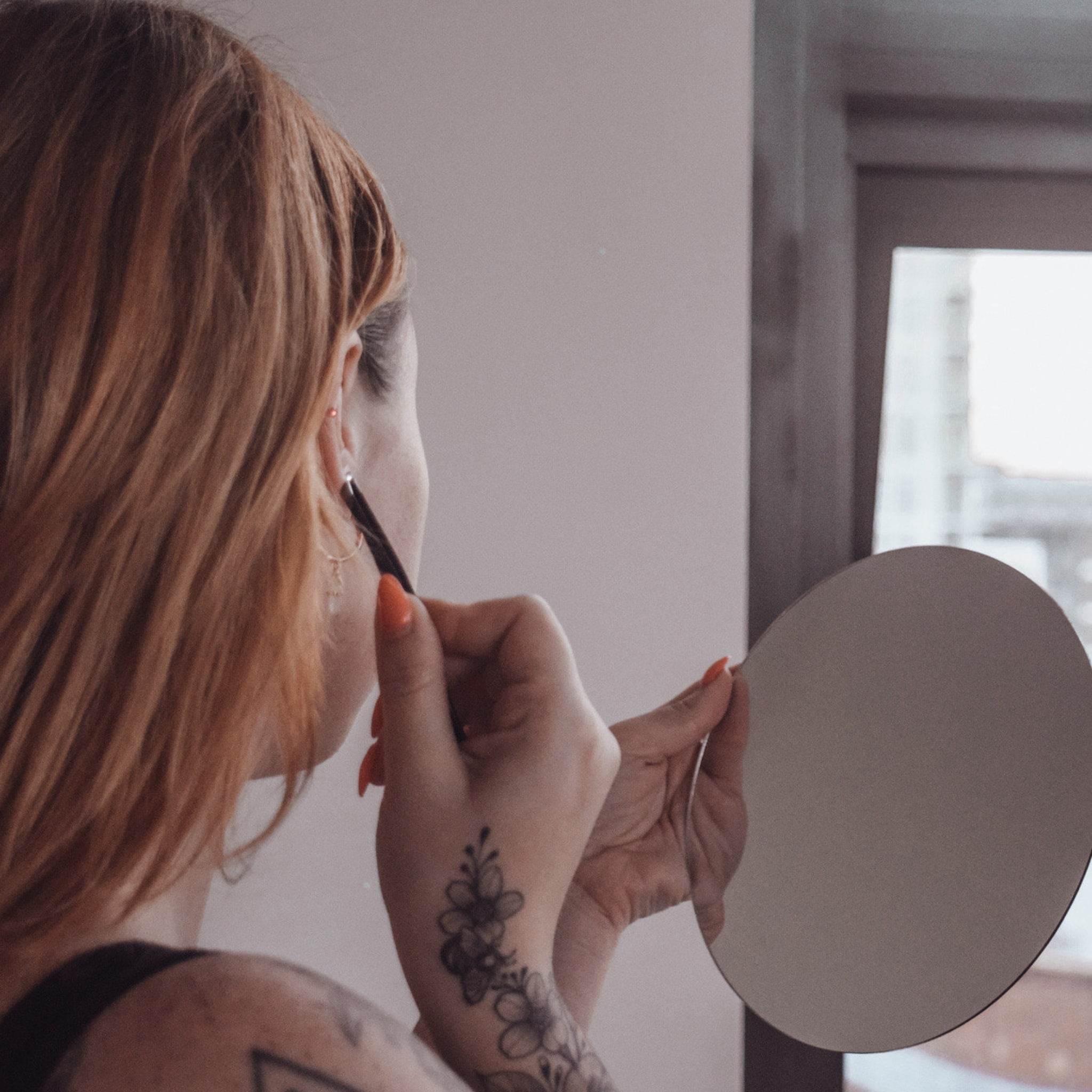 Woman holding mirror with red nails and floral tattoo, using tweezers