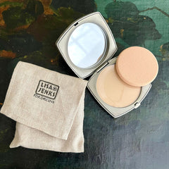 Powder compact with silver case, beige powder, and Lisa Jenks linen pouch on surface