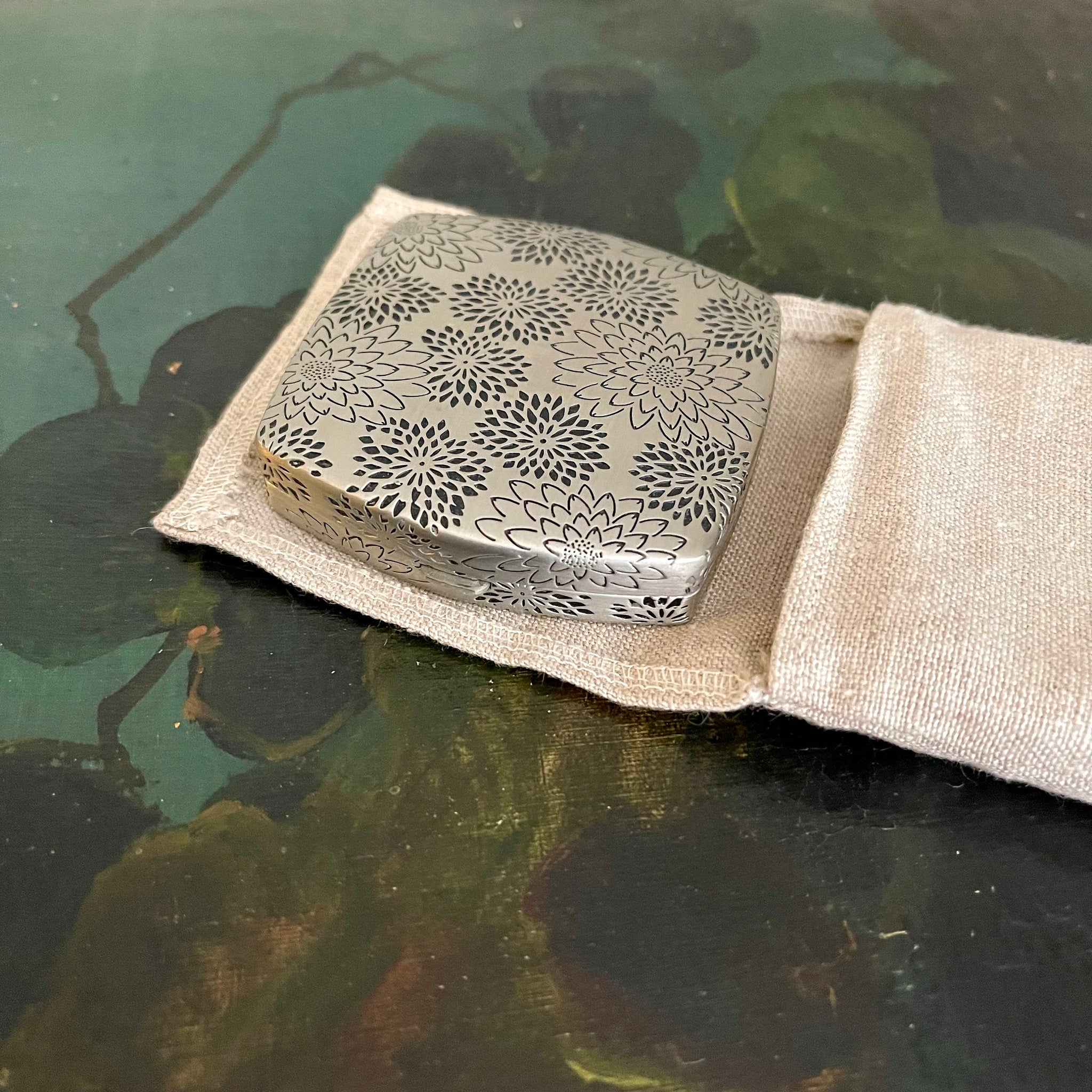 Metal floral patterned box in a beige linen pouch on a painted surface