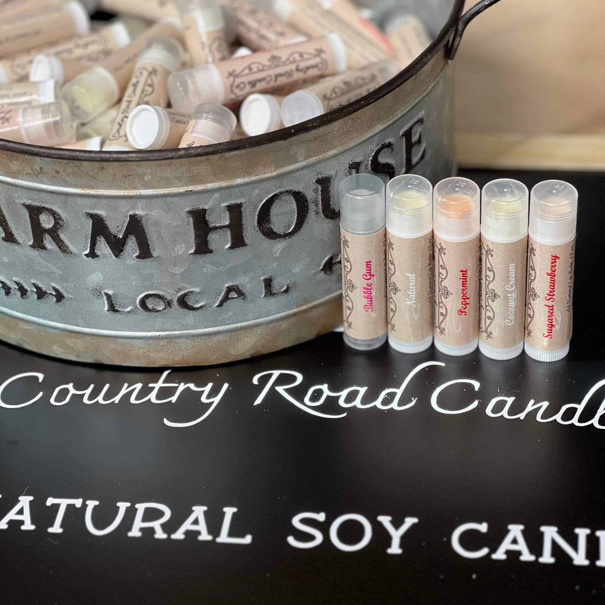 Lip balms in clear tubes with tan labels, Country Road Candle Co. branding visible, in a metal container