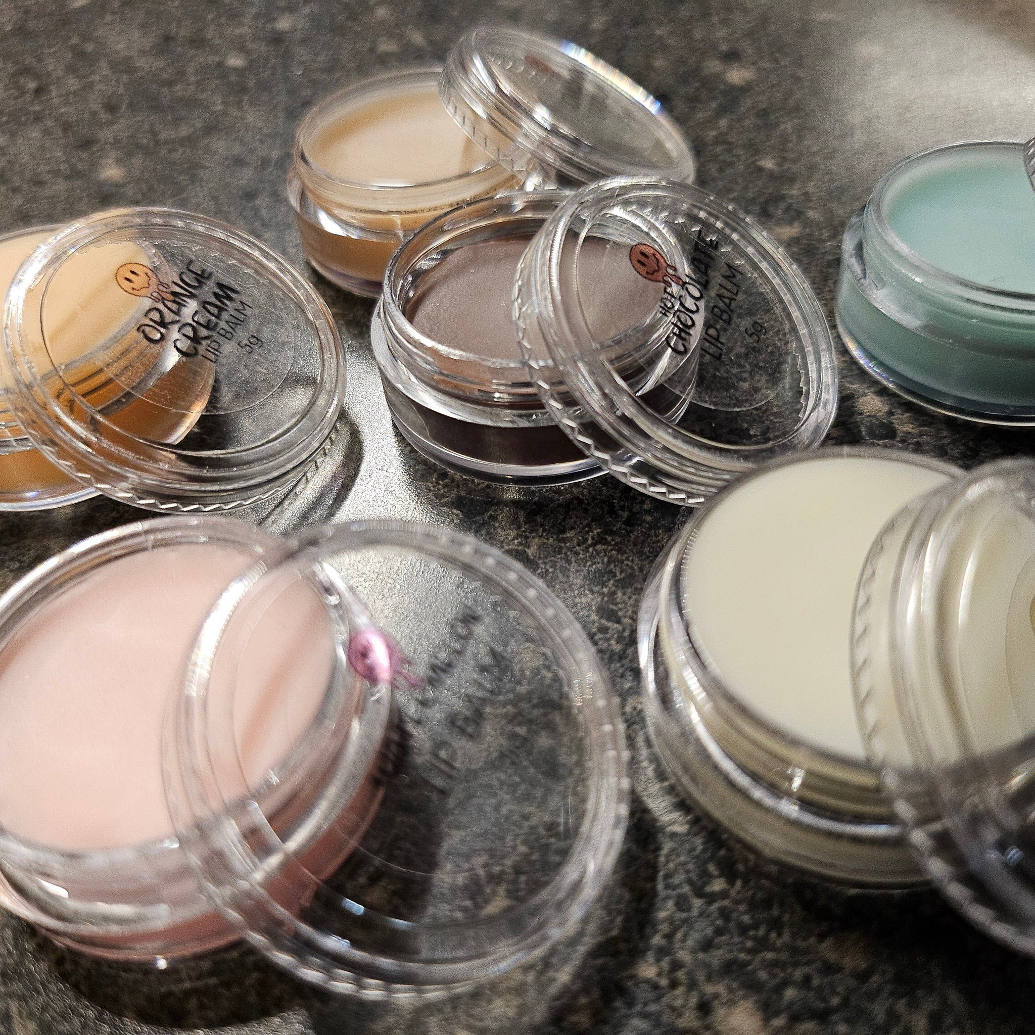 Lip balm pots with various colors in clear plastic containers, Orange Cream and Hot Choc labels visible