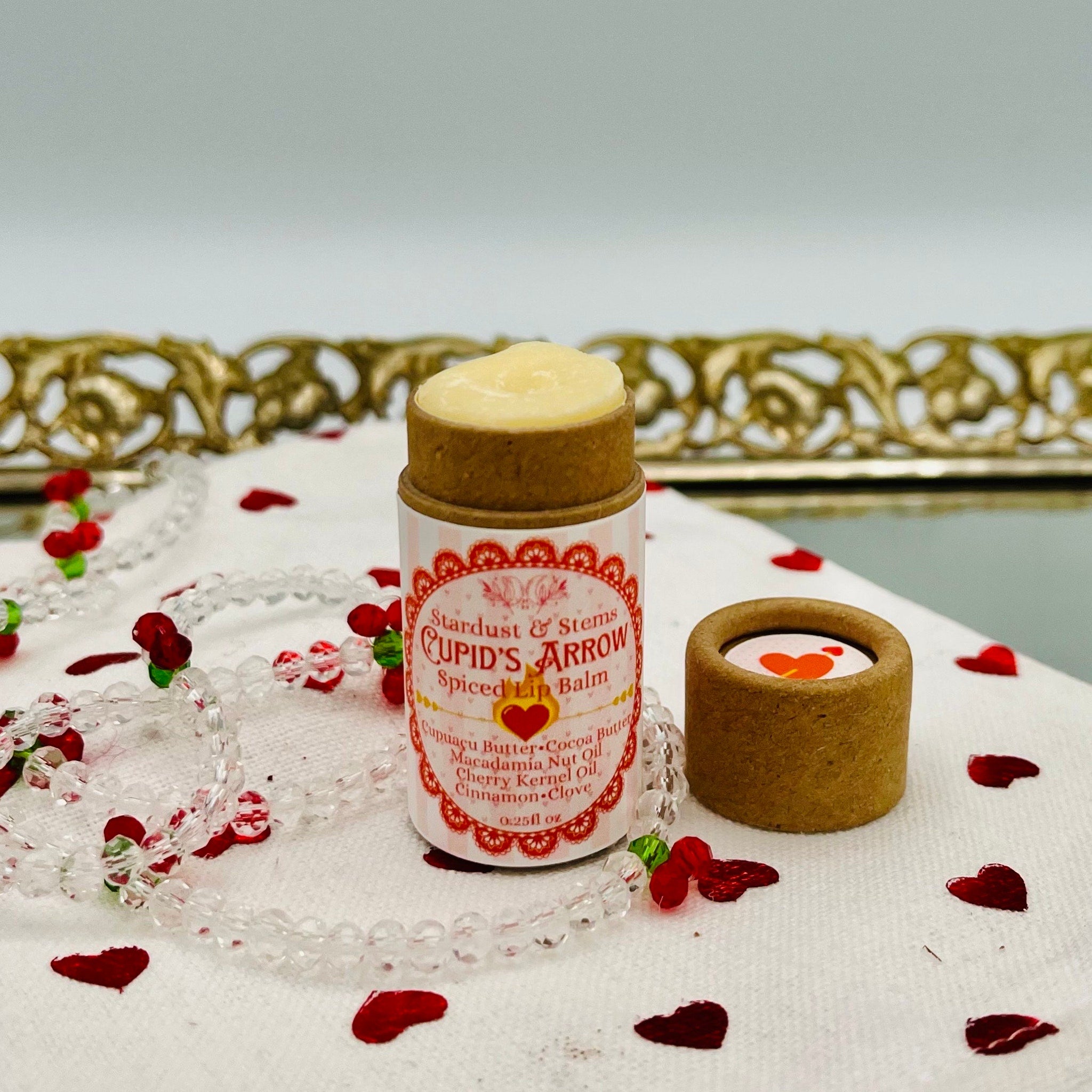 Lip balm tube with light pink stripes, brown cap, and "CUPID'S ARROW" label on white cloth with hearts