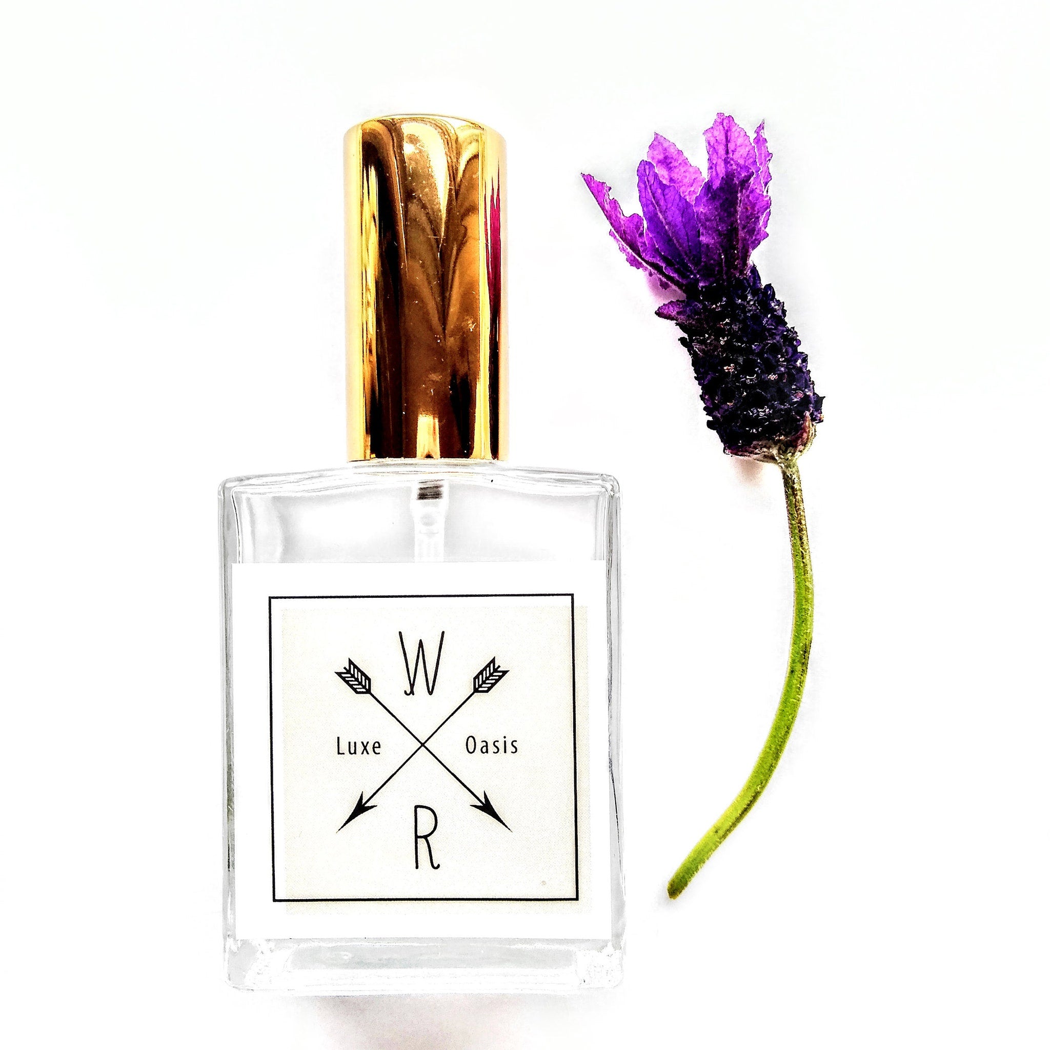 Perfume bottle with gold cap, clear glass, and Luxe Oasis label with purple flower next to it