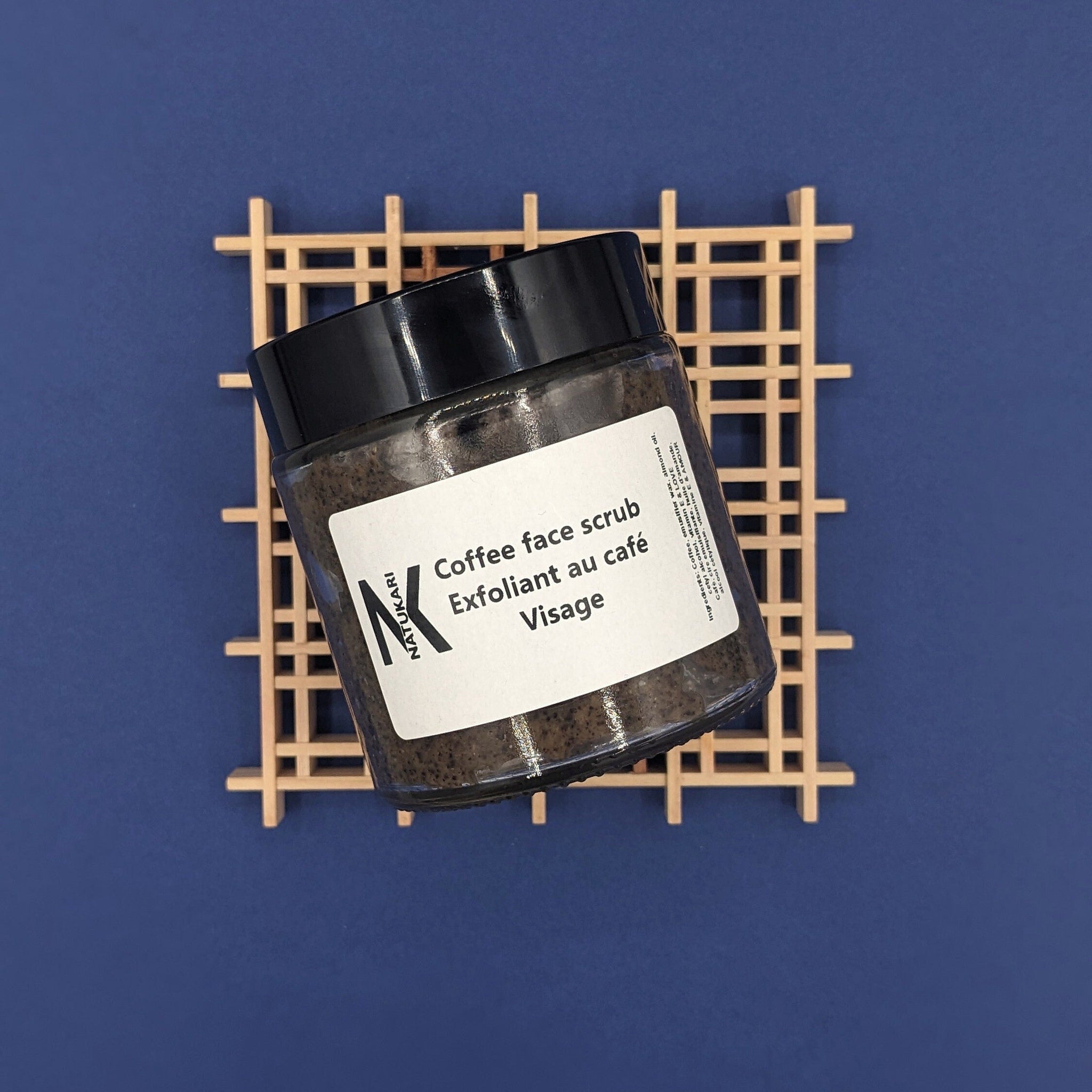 Coffee face scrub jar with black lid, visible granules, and NATUKARI label on a wooden grid