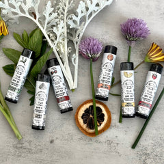 Lip balms with white tubes, black caps, and brand text, arranged with flowers and herbs