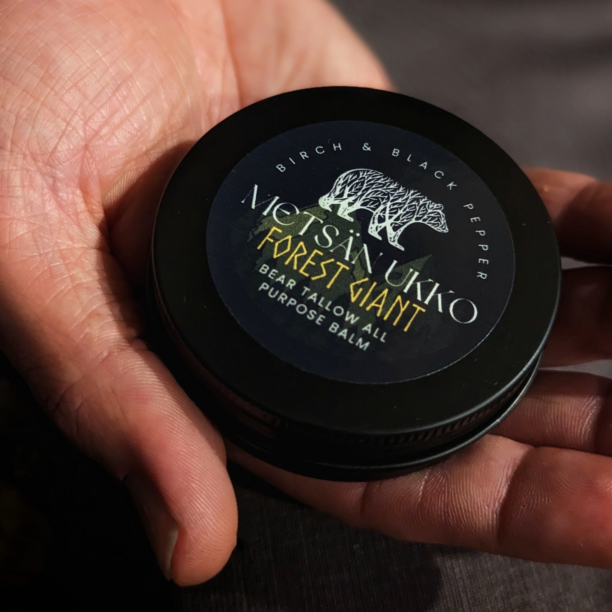 All purpose balm in black metal tin, held in hand, with Metsän Ukko Forest Giant label