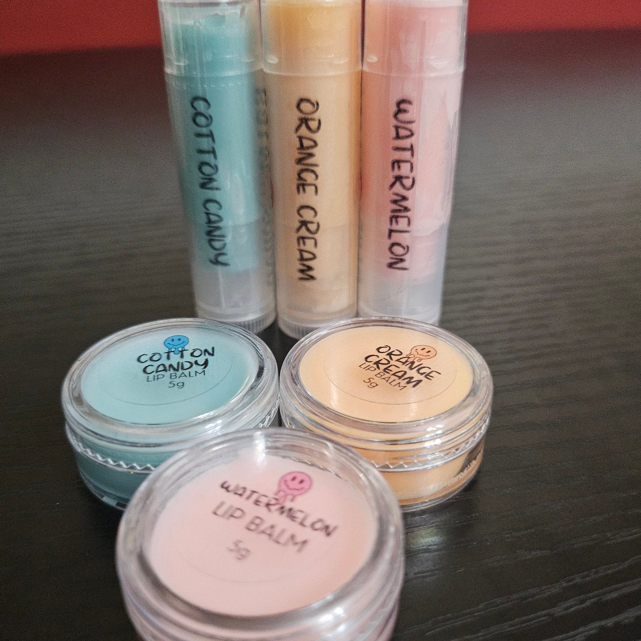 Lip balm tubes and jars in various colors, with handwritten labels: Cotton Candy, Orange Cream, Watermelon