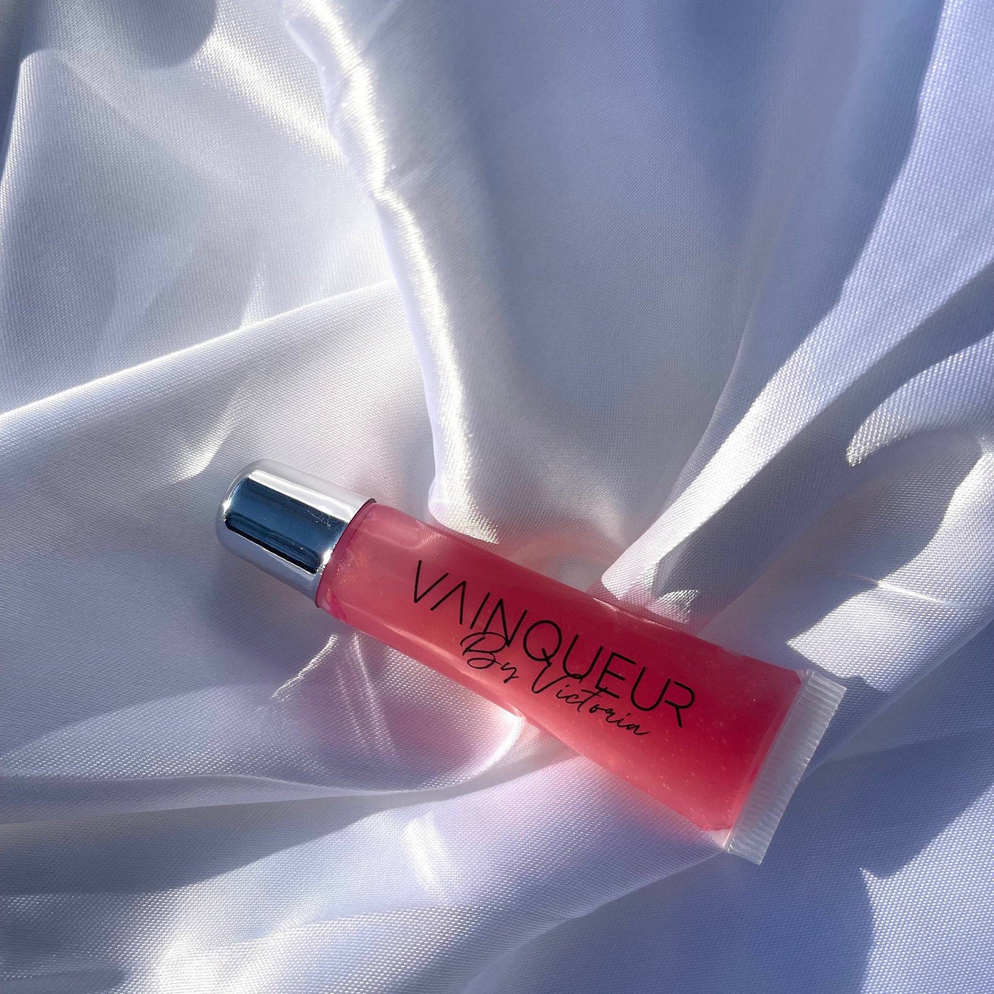 Lip gloss tube with pink gloss, silver cap, and Vainqueur Beauty text on white satin fabric