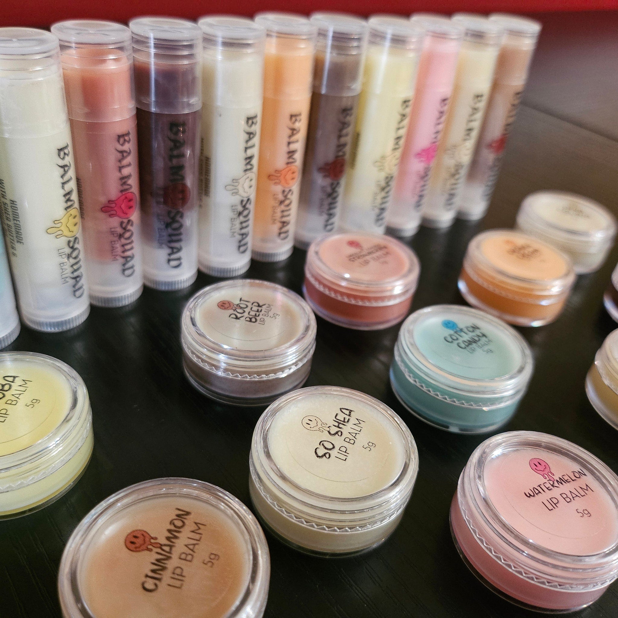 Lip balm tubes and jars in various colors, with "Balm Squad" and flavor labels visible on black surface