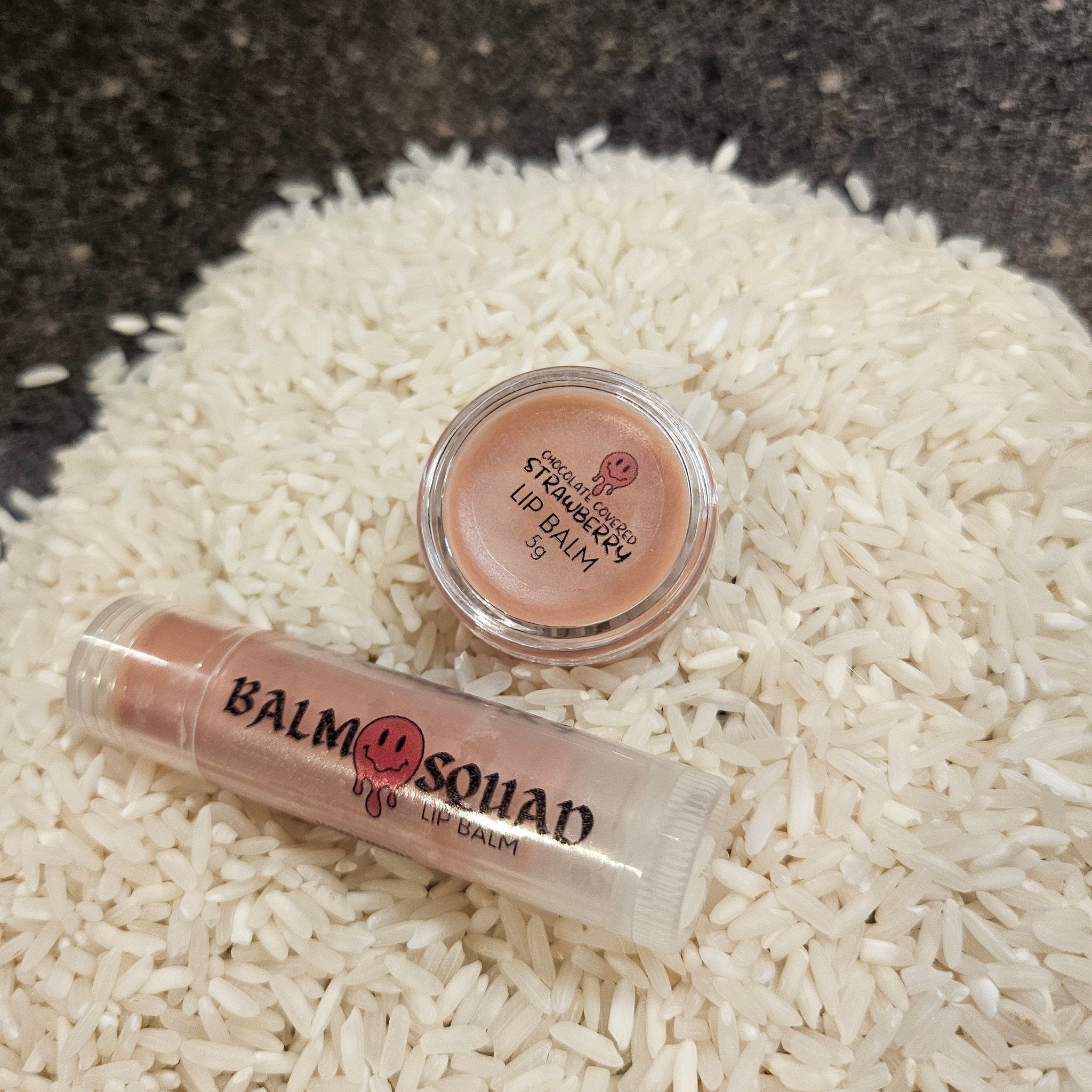 Lip balm tubes and jars in pink and clear plastic, on a bed of white rice, Balm Squad text visible