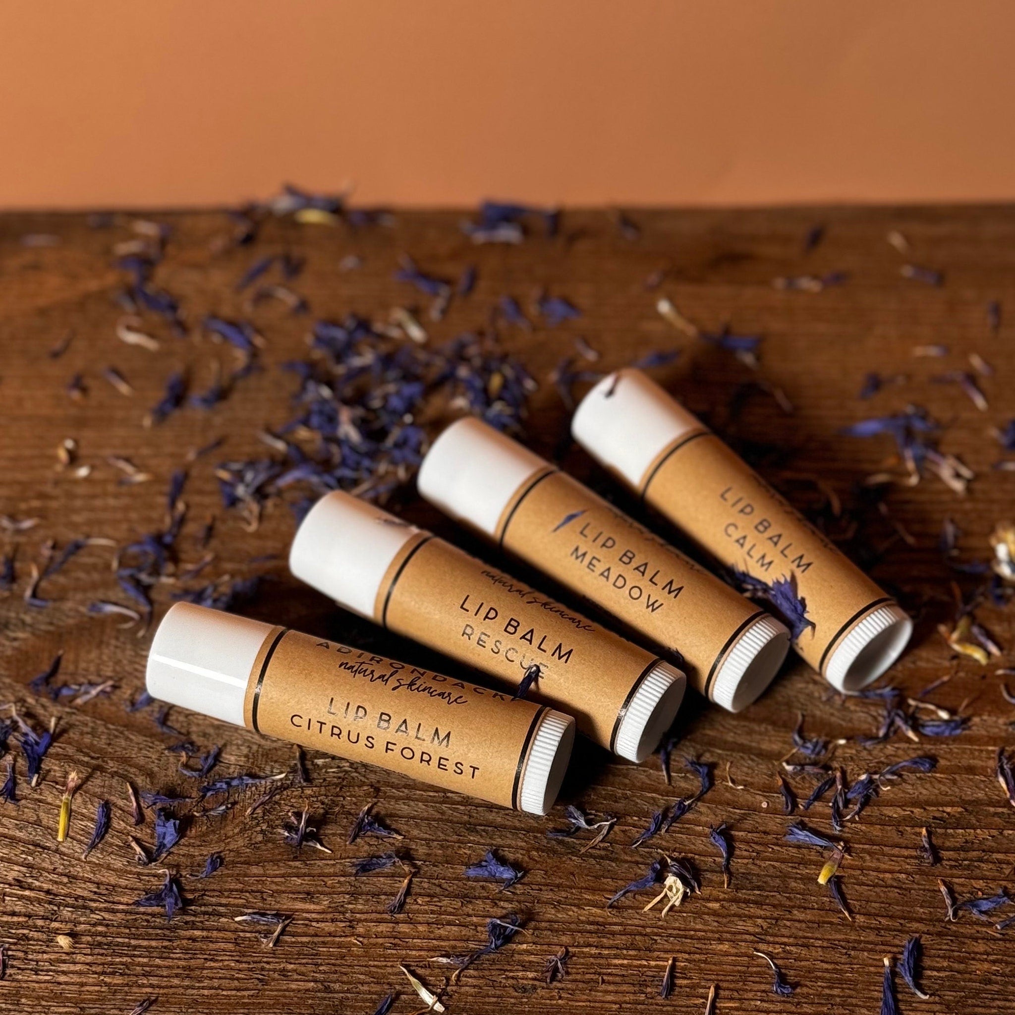 Lip balms with white caps, brown tubes, and text on a wooden surface with flower petals