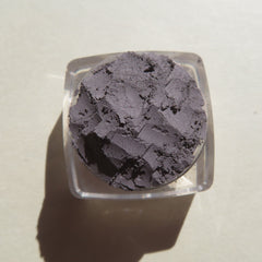 Powder eyeshadow in dark gray color with a rough texture in a clear plastic container