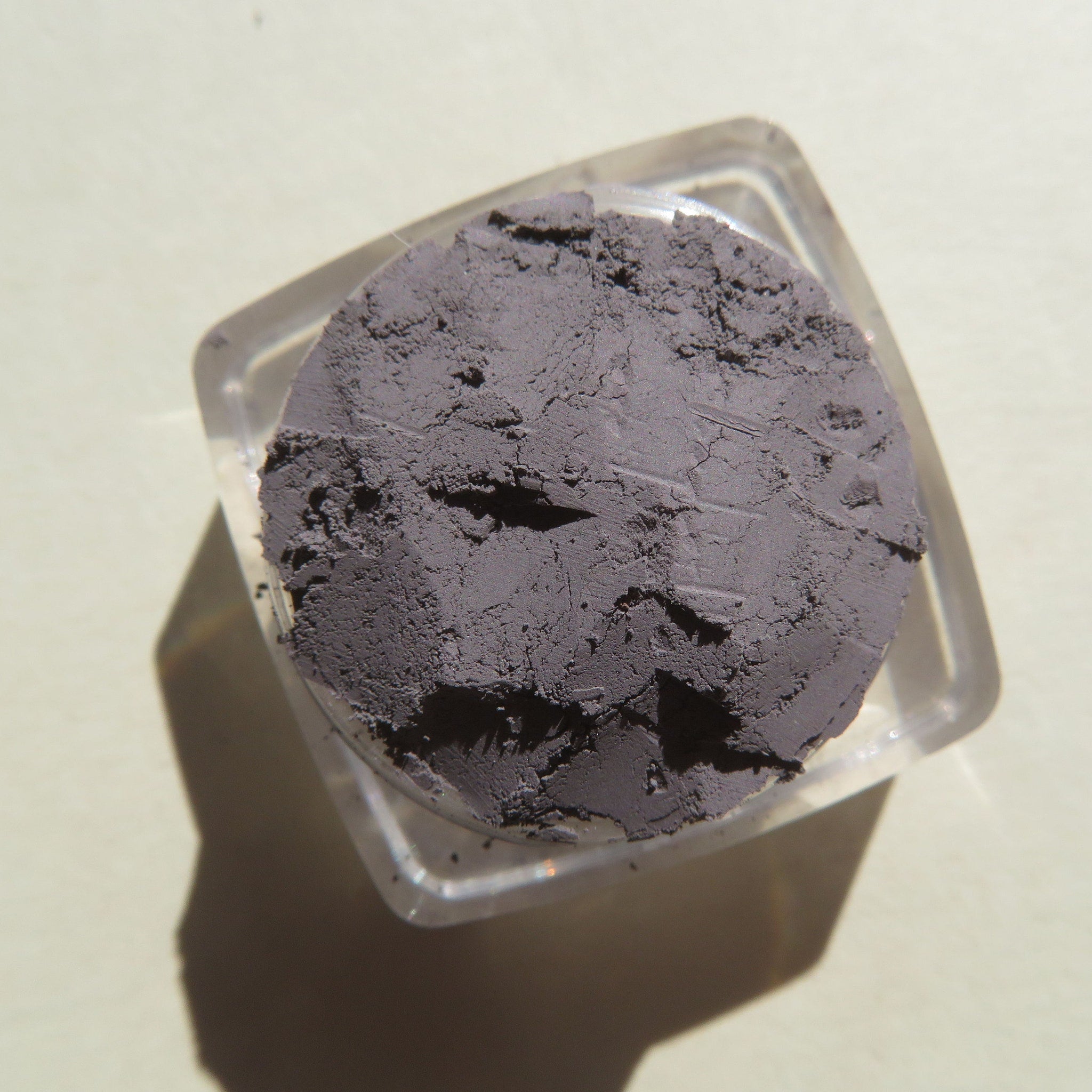 Powder eyeshadow in gray color with a cracked texture, inside a clear plastic container