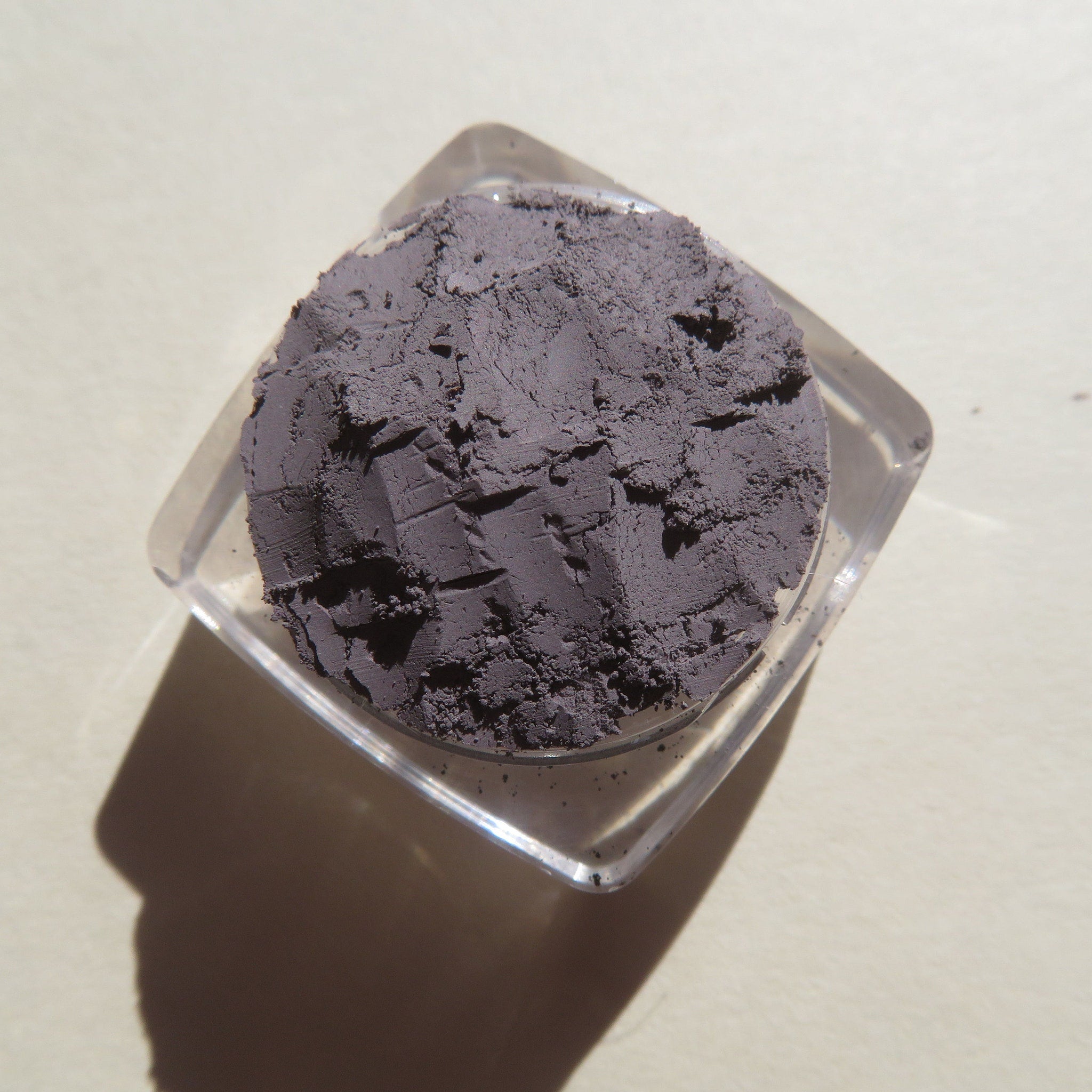 Powdered eyeshadow in dark gray, textured, in a clear plastic container, top view