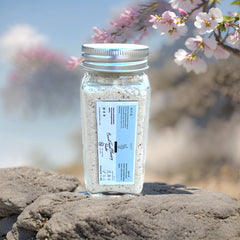 Facial Cleansing Powder in a clear glass jar with a silver lid, on a rock, with pink flowers in background