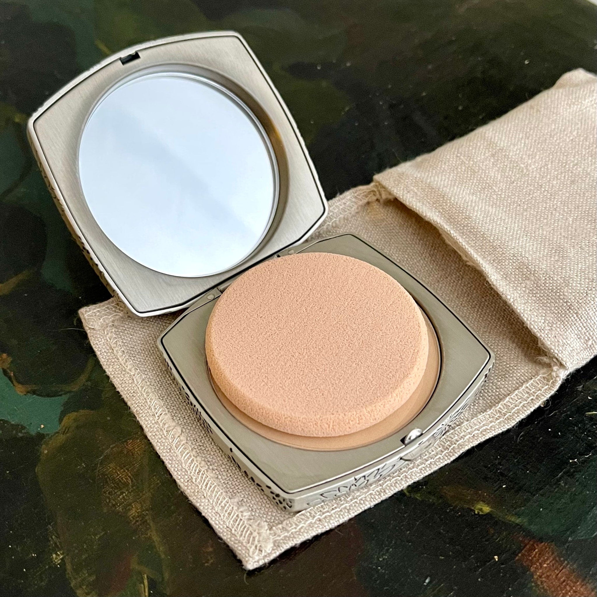 Compact powder with silver metal case, beige sponge, and linen pouch on a dark surface