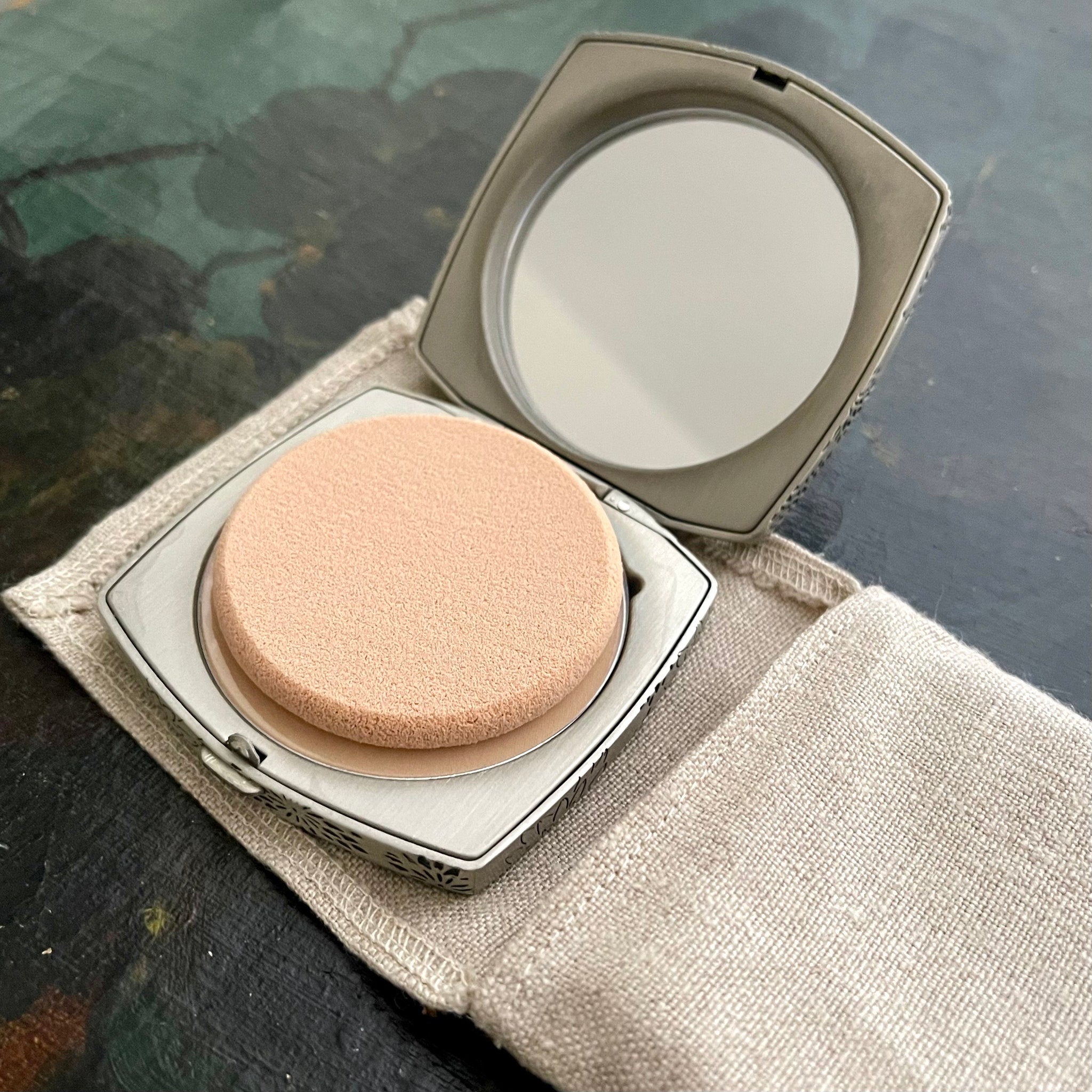 Compact powder with beige sponge, silver metal case, and linen pouch on table