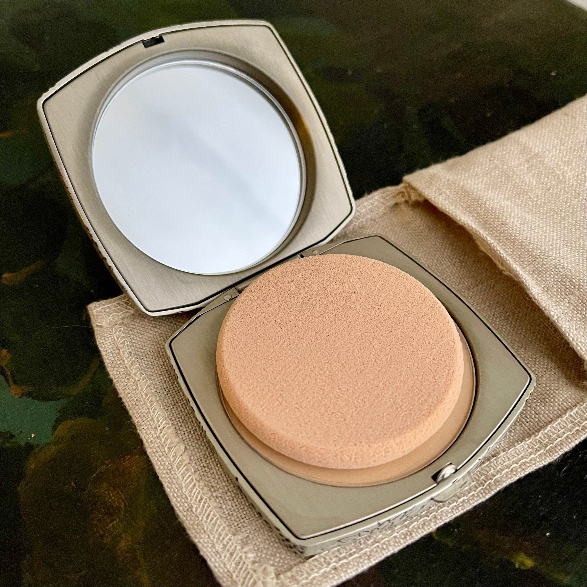 Compact mirror with silver metal case, beige sponge, and linen pouch