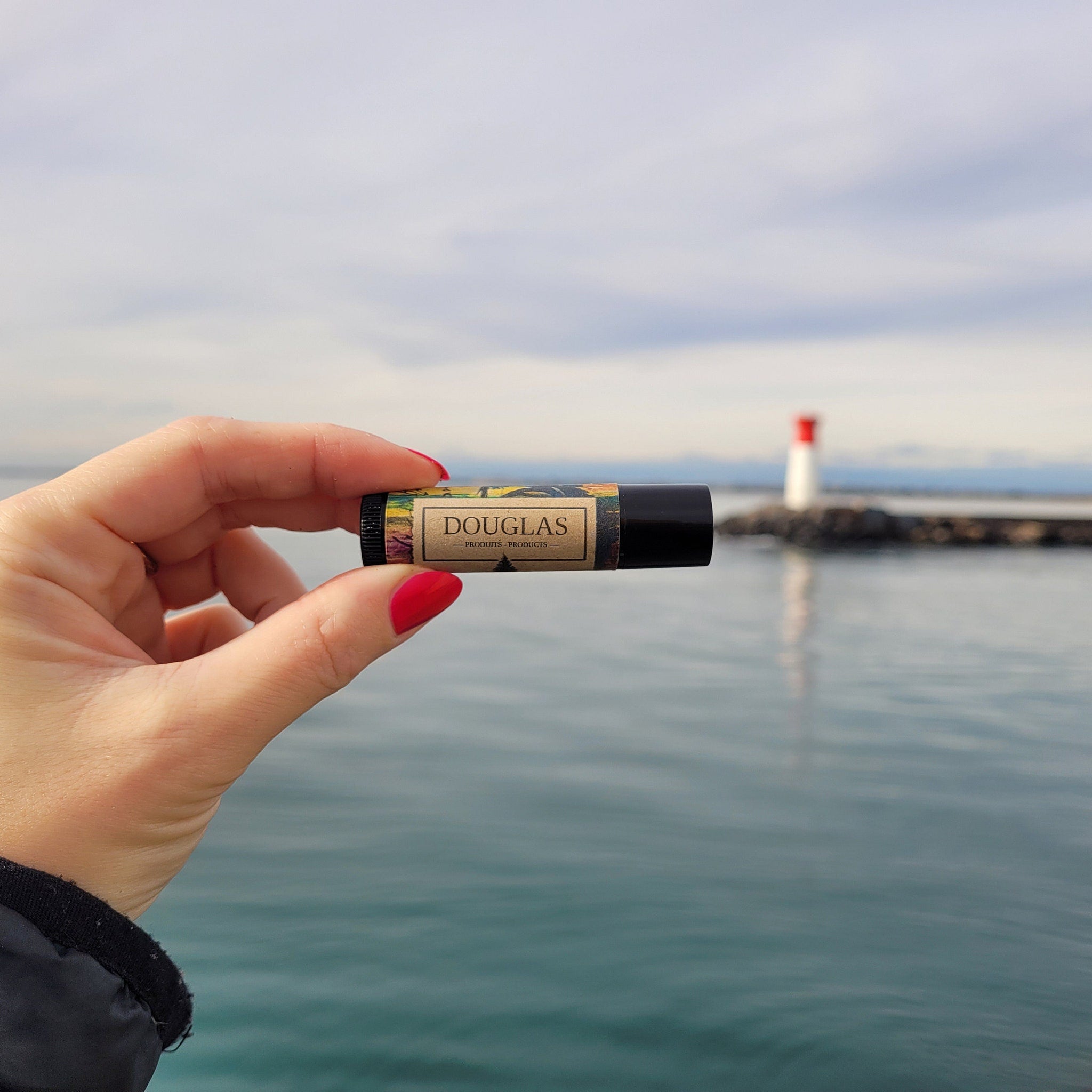 Lip balm tube with black cap, printed label, and DOUGLAS branding held in hand near water