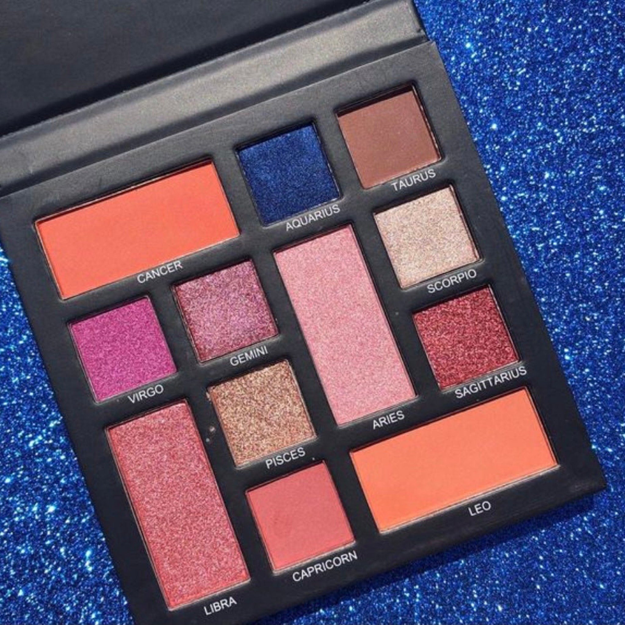Eyeshadow palette with multiple colors, glittery finish, and zodiac labels in a black case