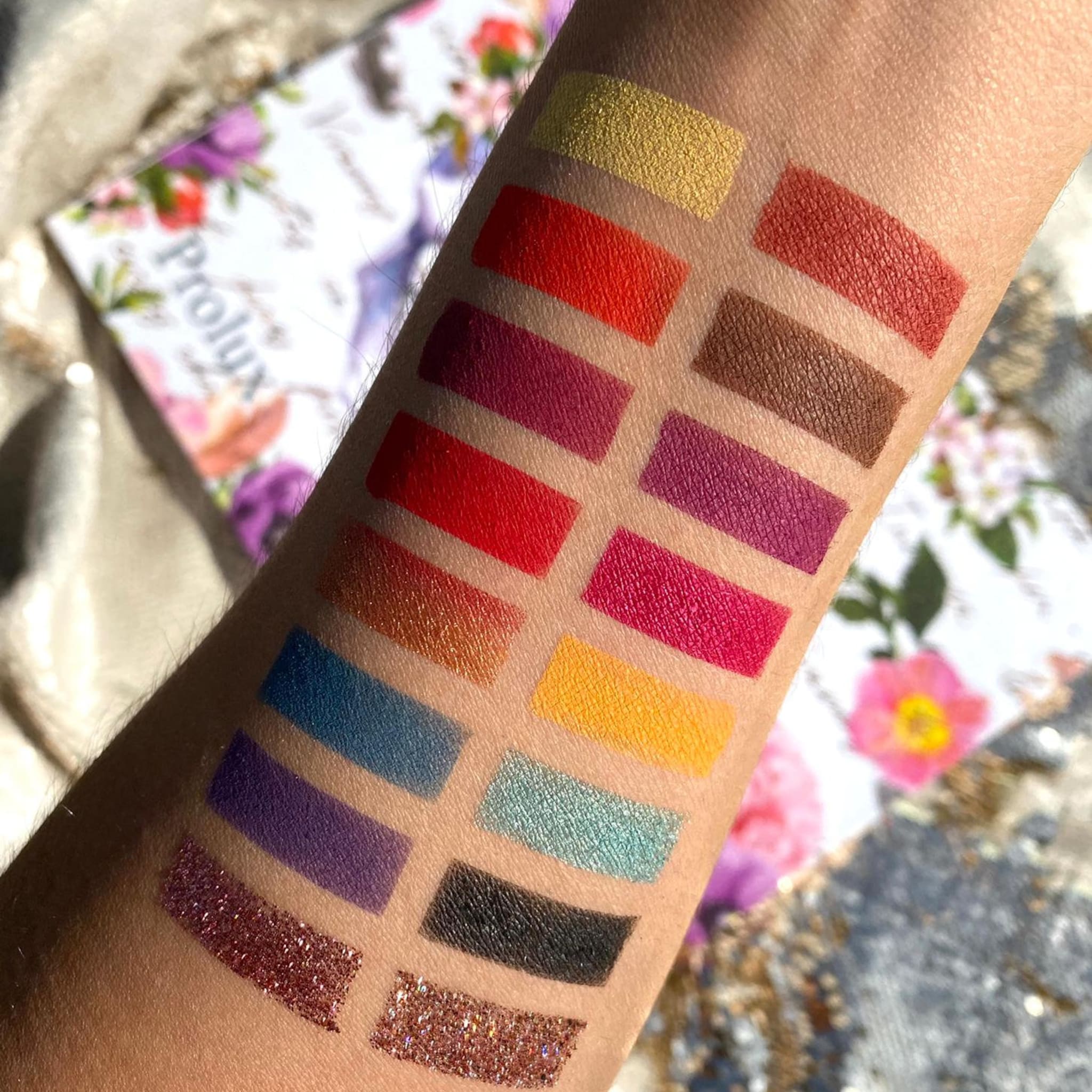 Eyeshadow swatches, various colors including red, gold, and purple, on arm, Prolux brand visible