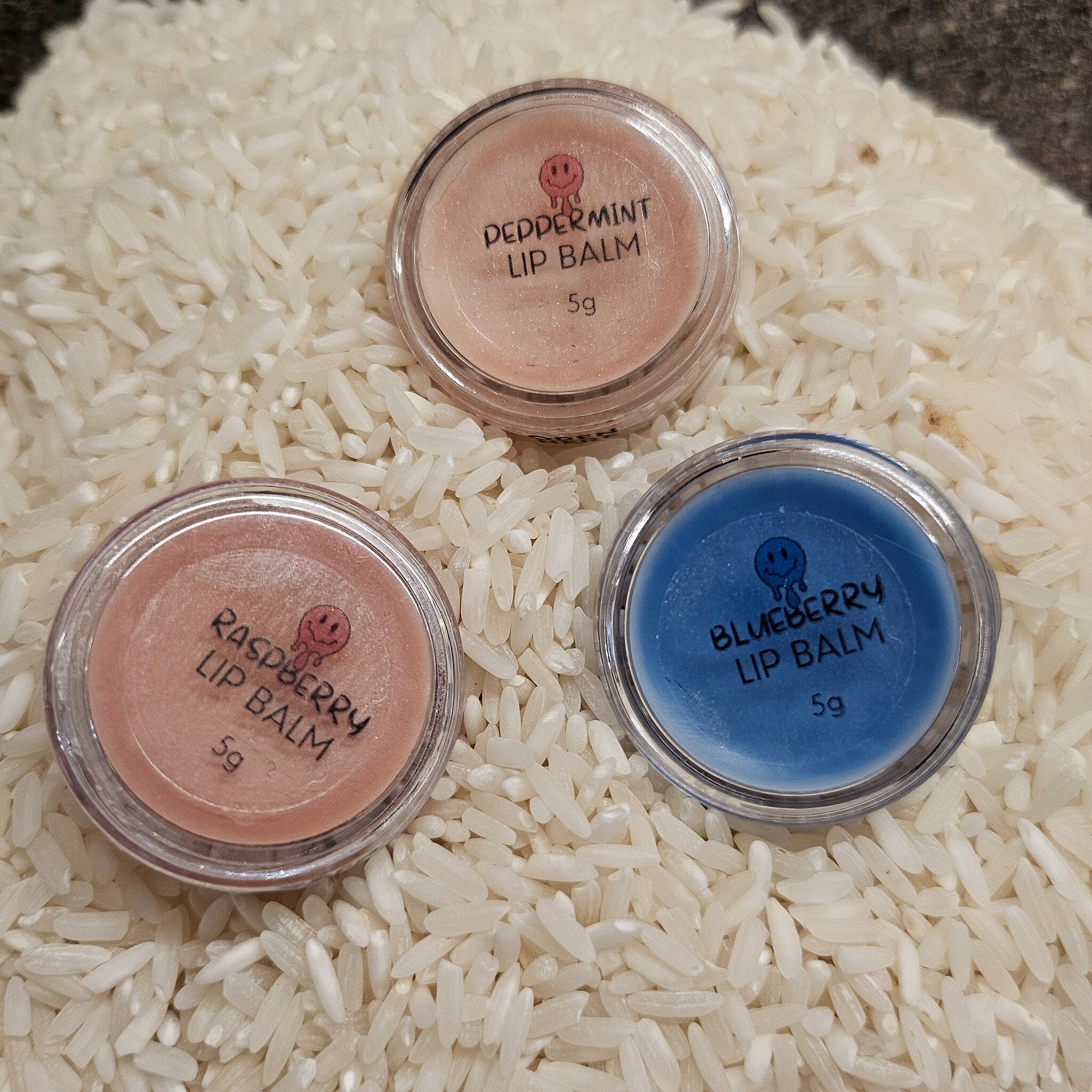 Lip balm pots, clear plastic, pink, blue, and peach colored contents, on white rice bed