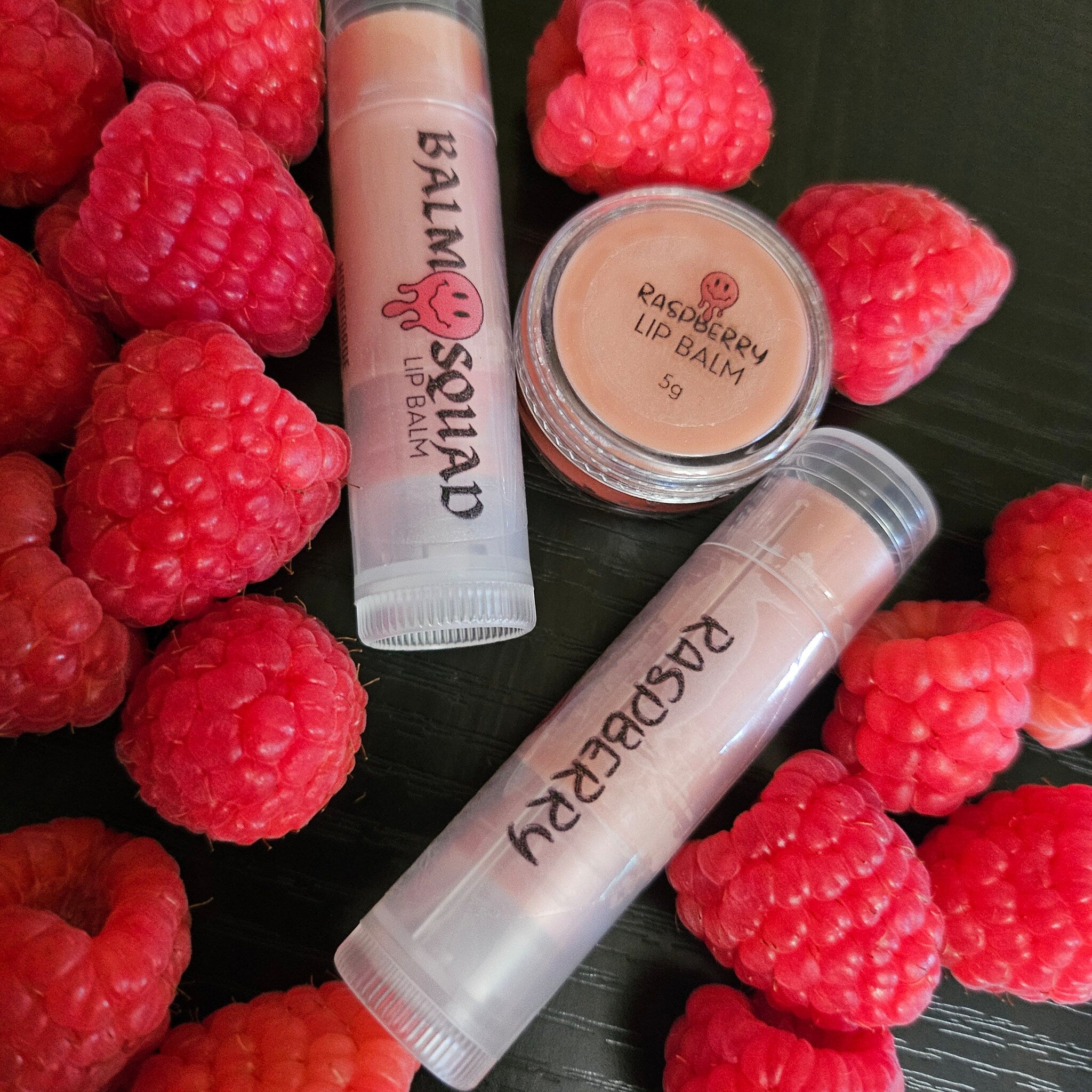 Lip balm tubes and jar with pink balm, clear tubes, and red raspberries on dark surface